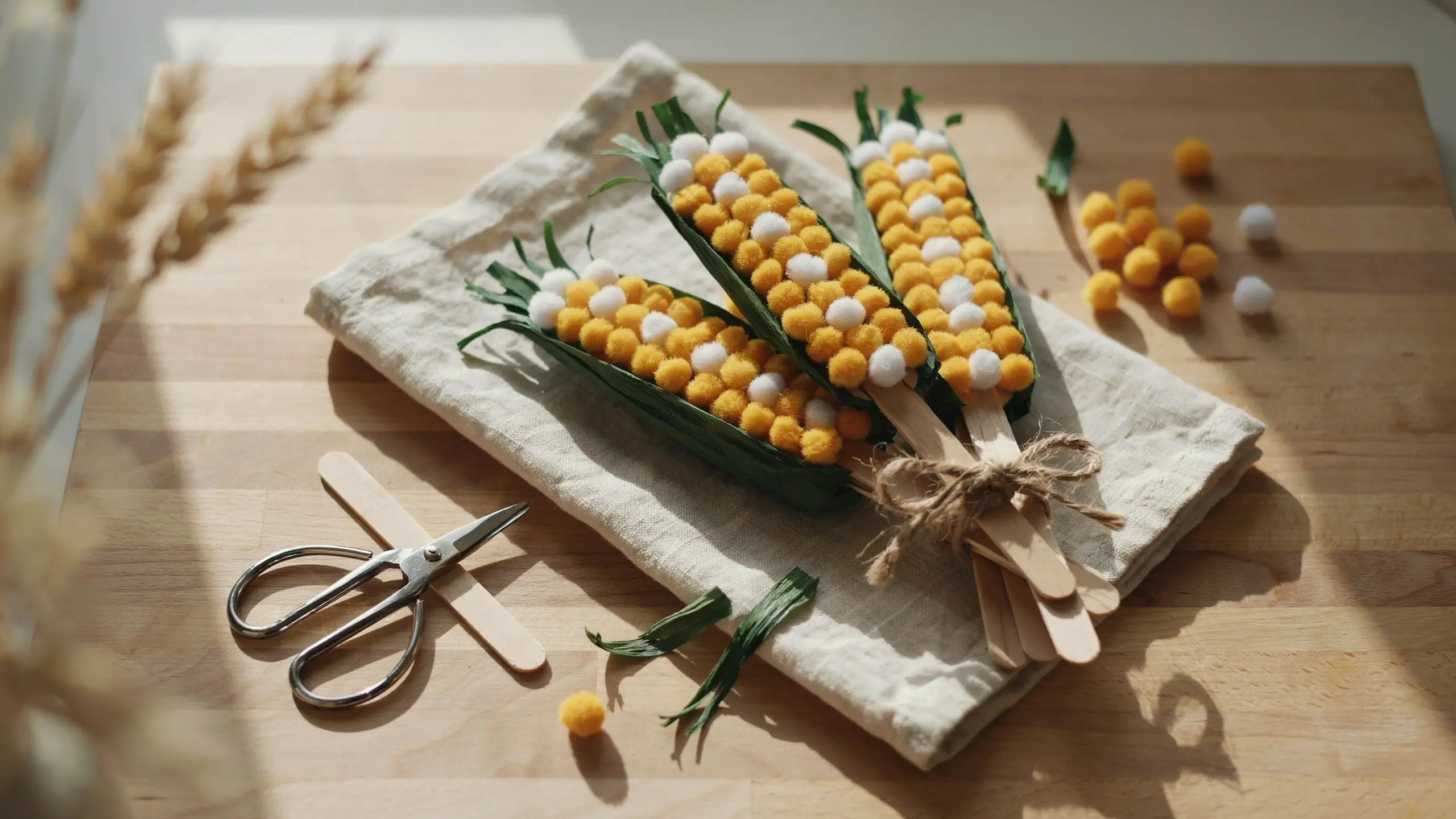 Close-up of a DIY popsicle stick corn craft featuring yellow and white pom-pom kernels and green paper husks tied with rustic twine on a wooden table