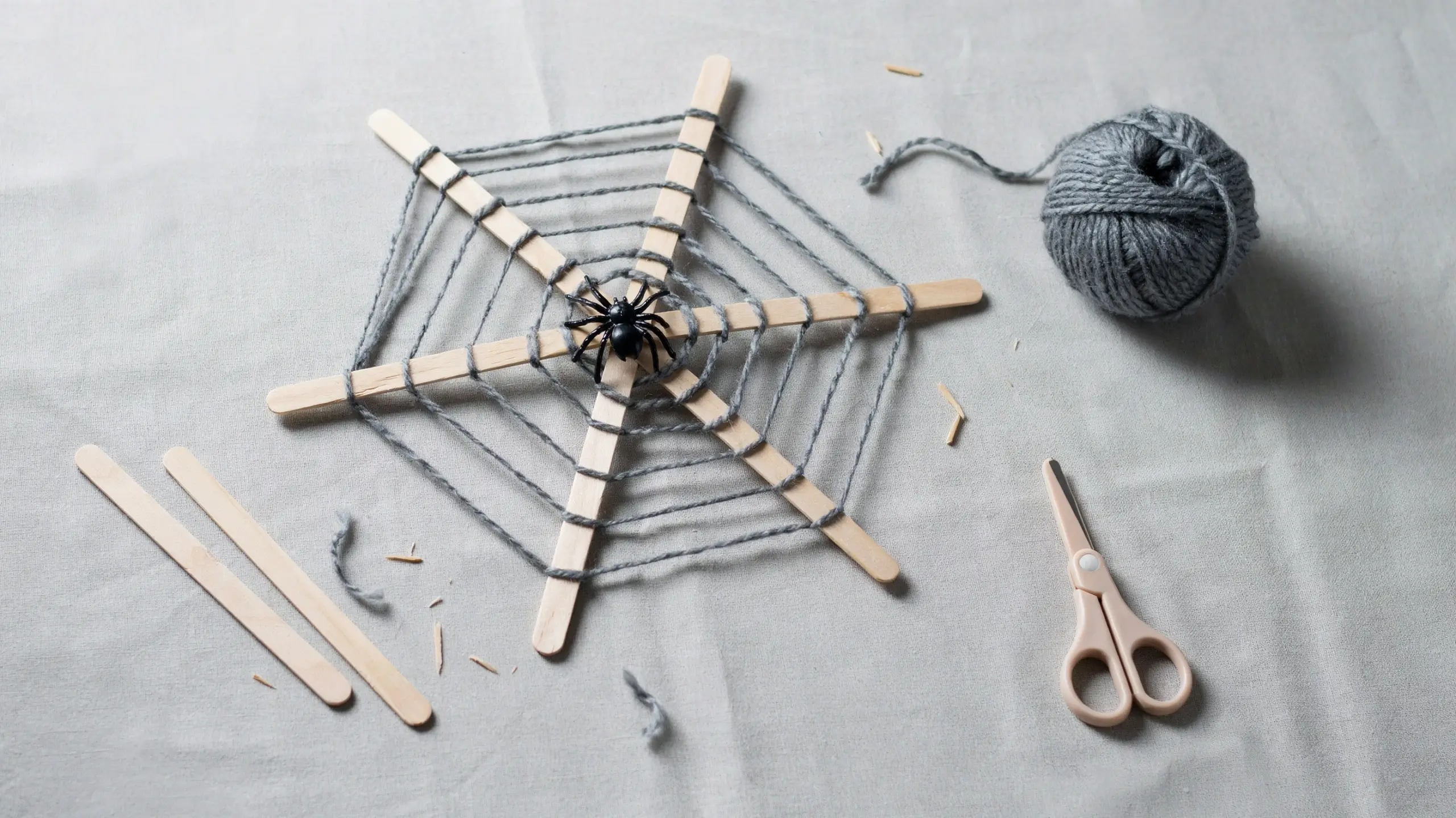 DIY popsicle stick spiderweb craft with grey yarn weaving and a fake black spider on a textured background