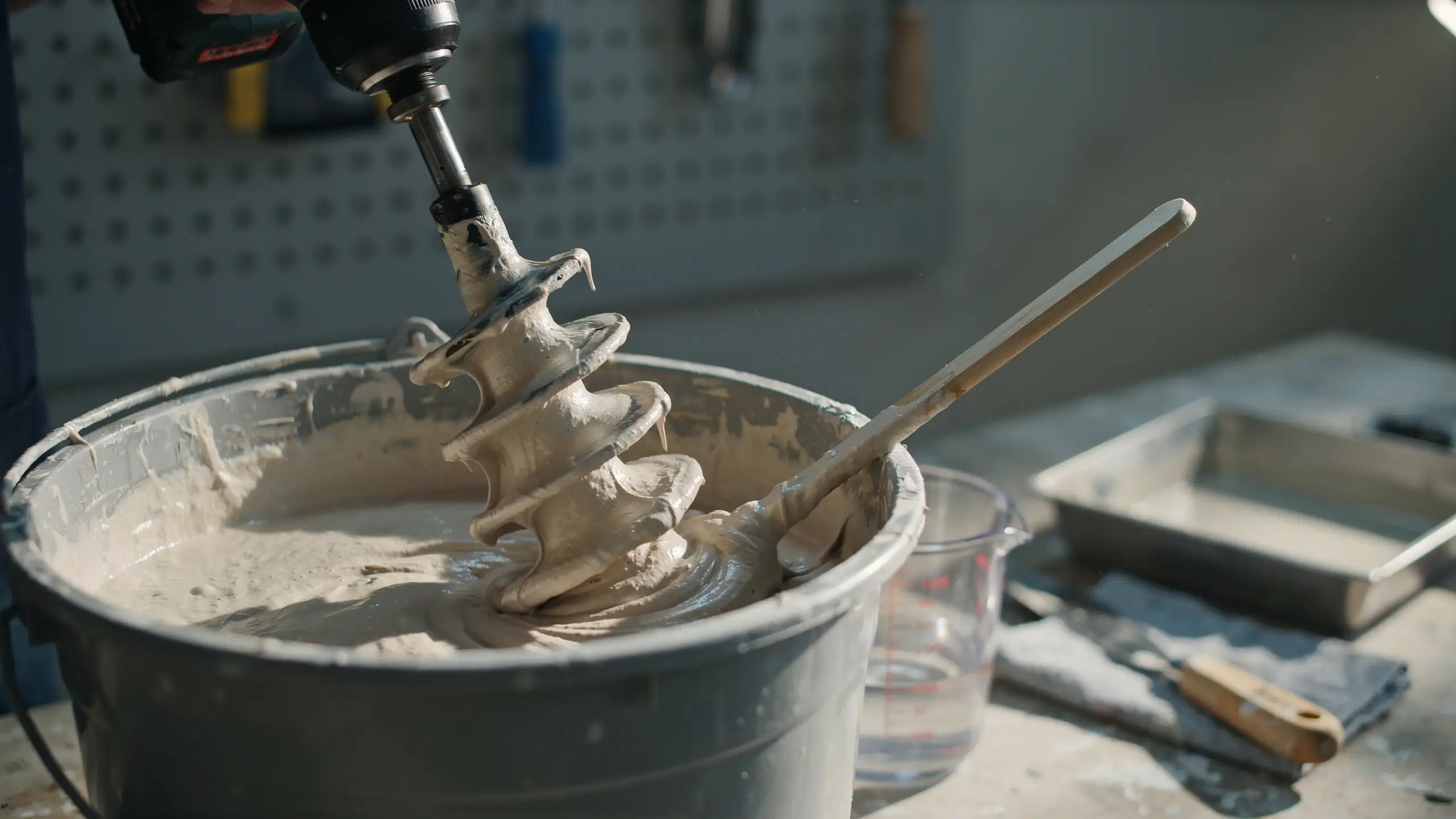 A close-up shot of a power drill with a spiral mixing paddle thoroughly stirring thick, white drywall joint compound inside a grey plastic bucket on a workshop workbench.