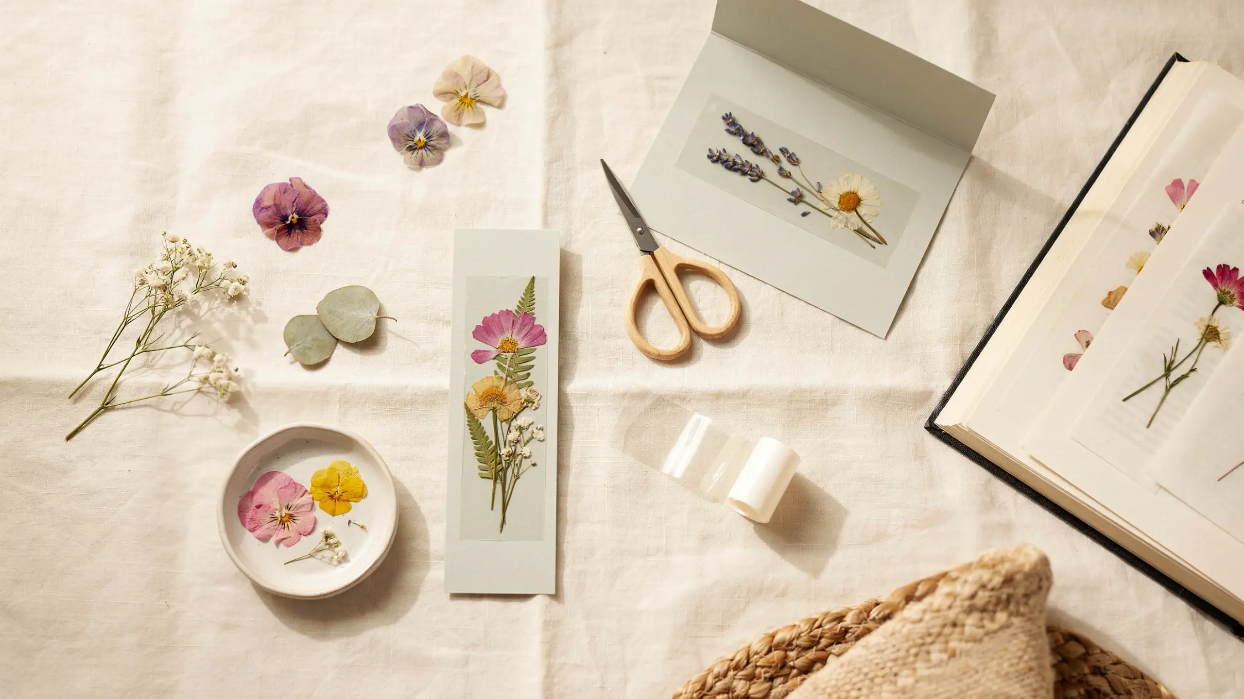Pressed flower bookmark and card craft with dried flowers, scissors, glue, and an open flower-pressing book.