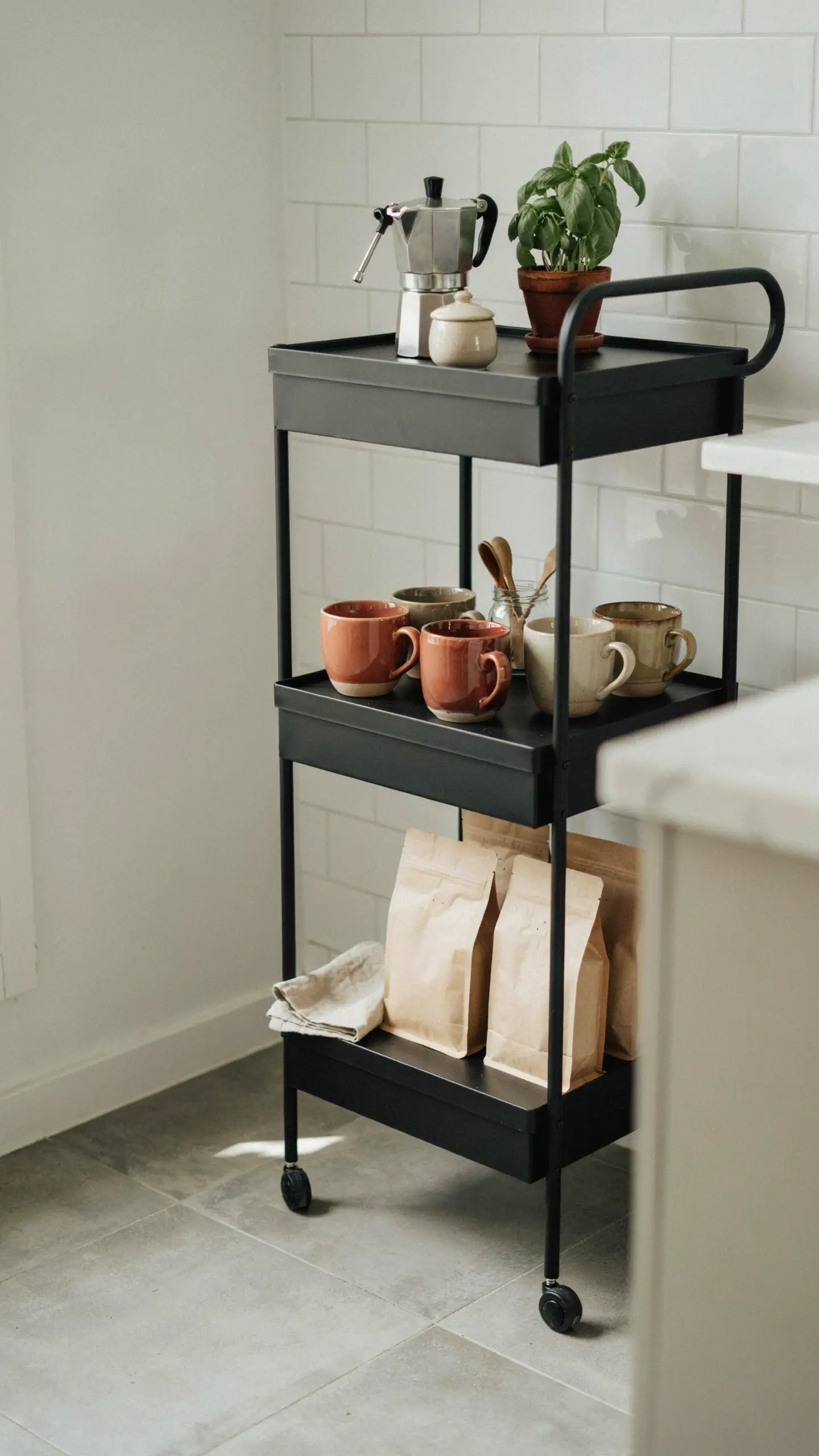 Black IKEA RASKOG utility cart styled as a compact coffee station, holding a moka pot, a potted basil plant, ceramic mugs, and brown kraft bags of coffee beans against a white subway tile backsplash. 