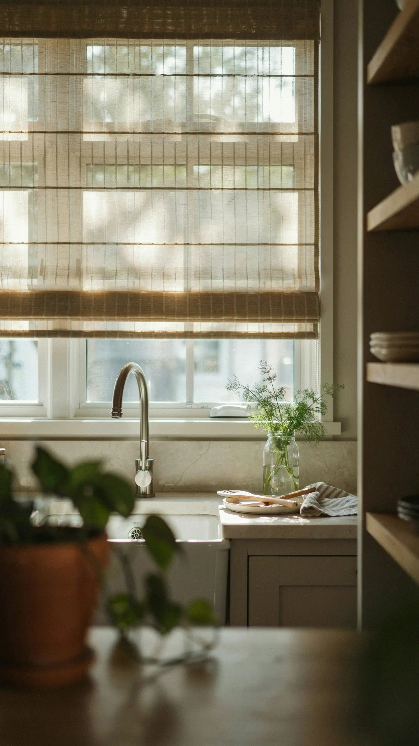Rattan Roman shade over a kitchen sink window with warm sunlight, greenery, and a farmhouse sink.