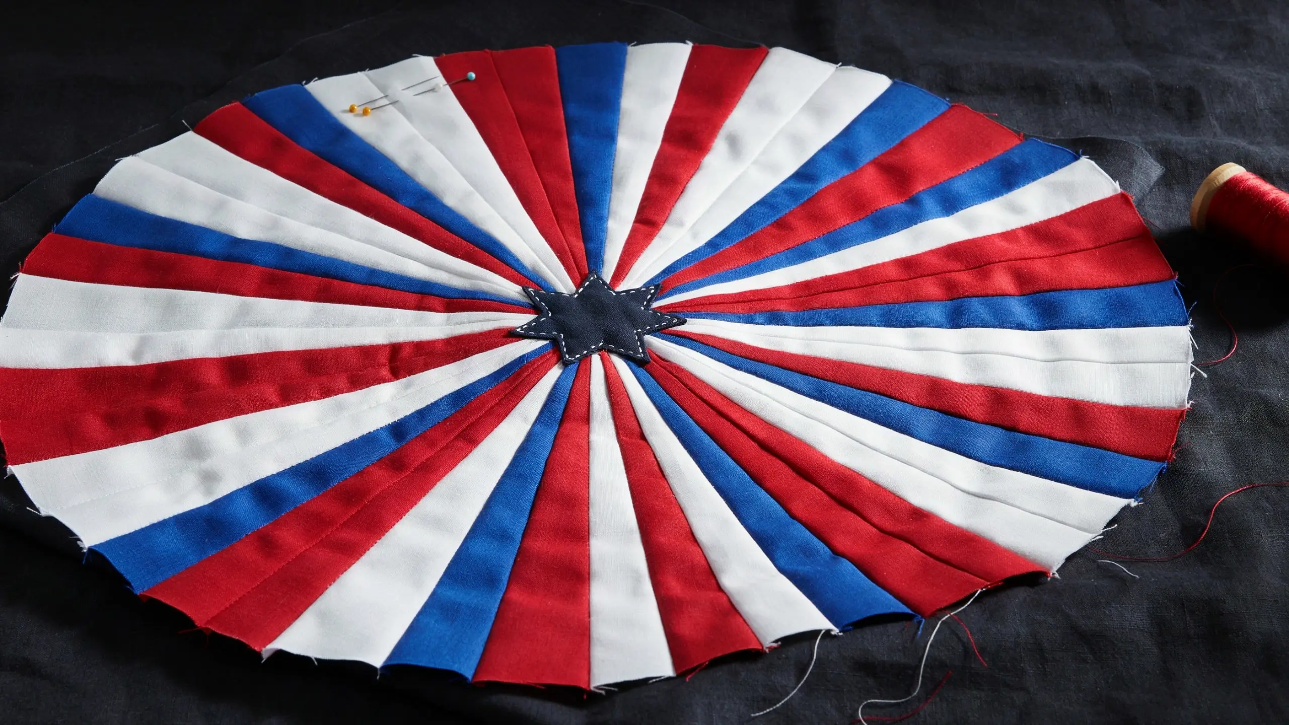 Top-down view of a red, white, and blue radiating stripe quilt patchwork block with a dark star center, pinned to a black fabric background next to a red thread spool, representing a fireworks burst design.