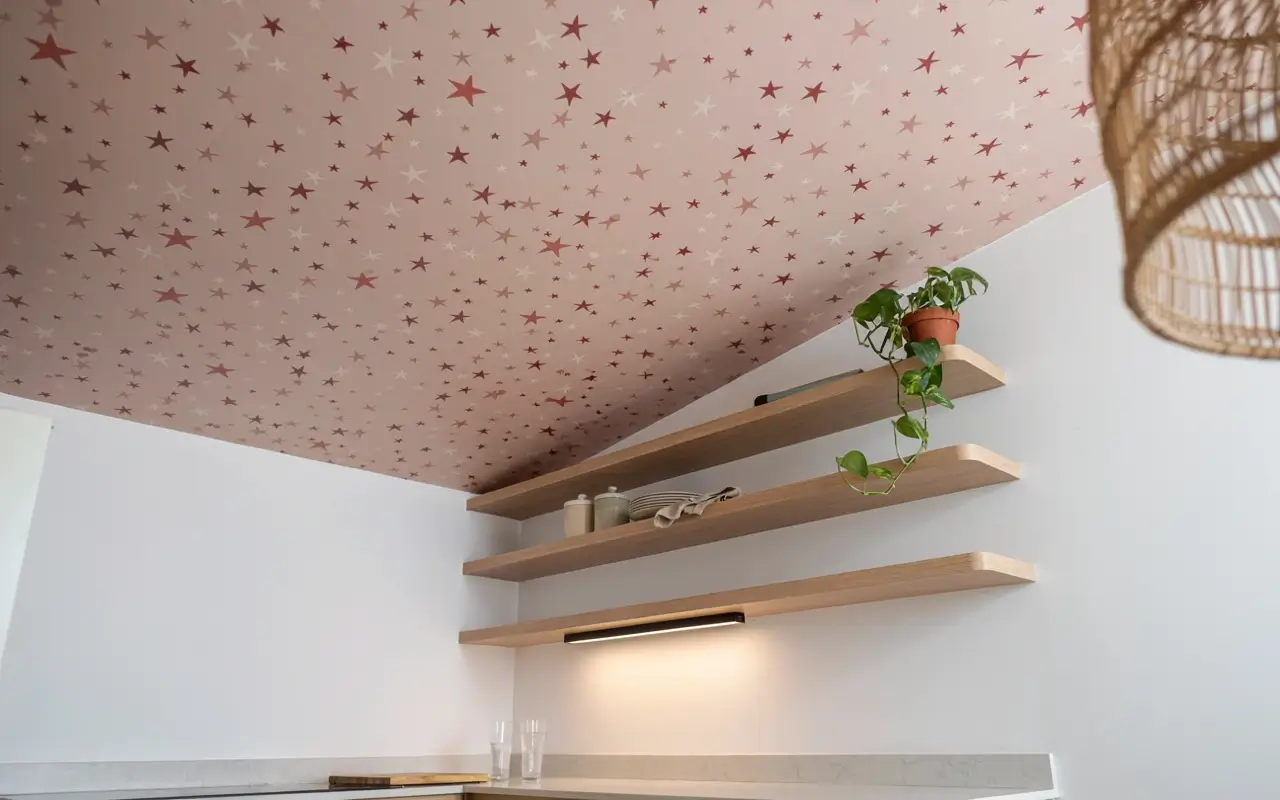 Kitchenette niche ceiling covered with removable pink star wallpaper above open wood shelves.
