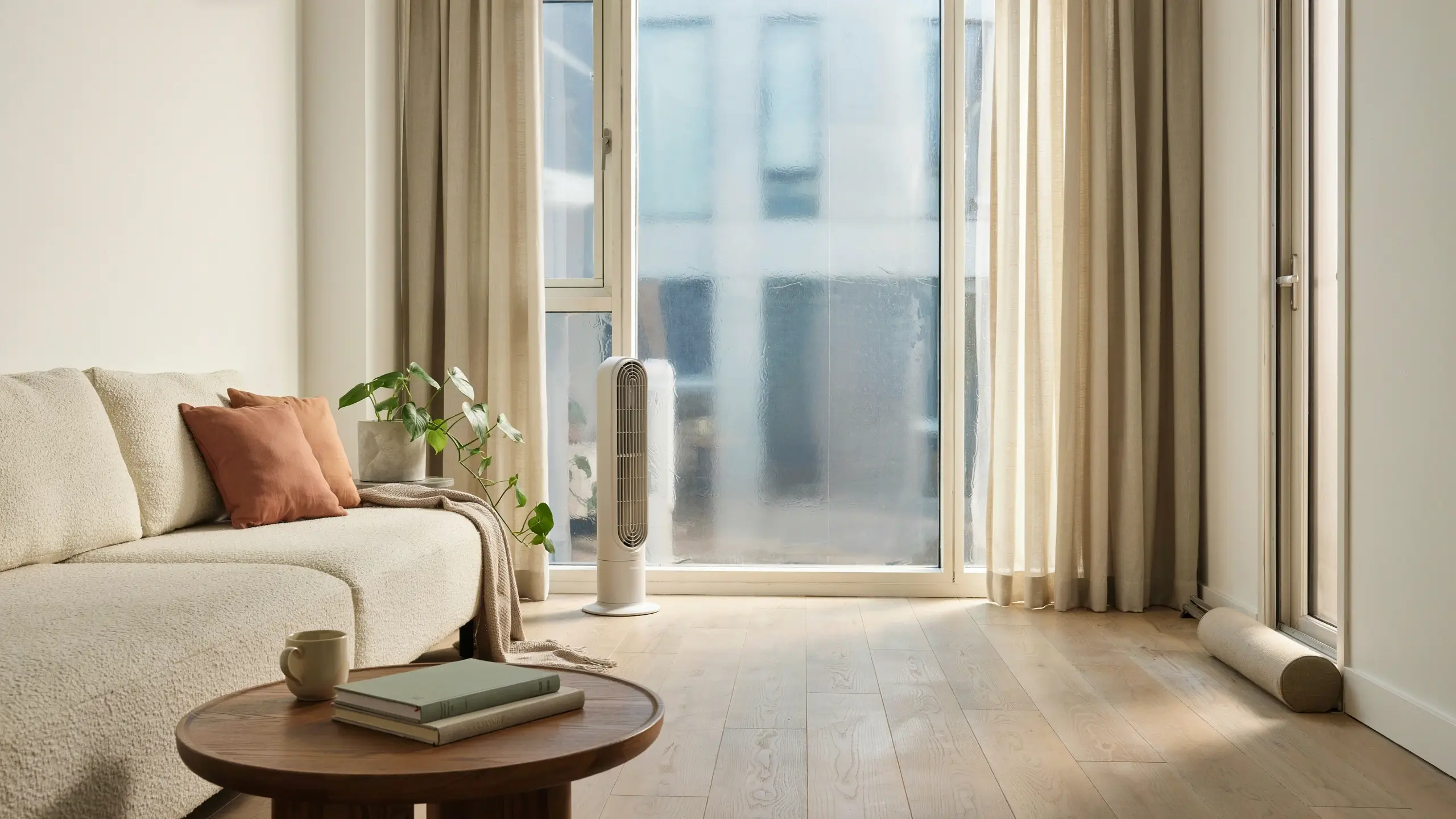 A cozy, renter-friendly living room featuring a white boucle sofa with rust-colored throw pillows, a white tower fan, and a round wooden coffee table, set against a large sliding glass door covered in removable frosted privacy film.