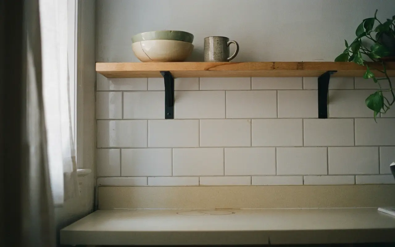 Wood kitchen shelf on renter-safe black brackets above white subway tile with bowls, mug, and trailing plant.