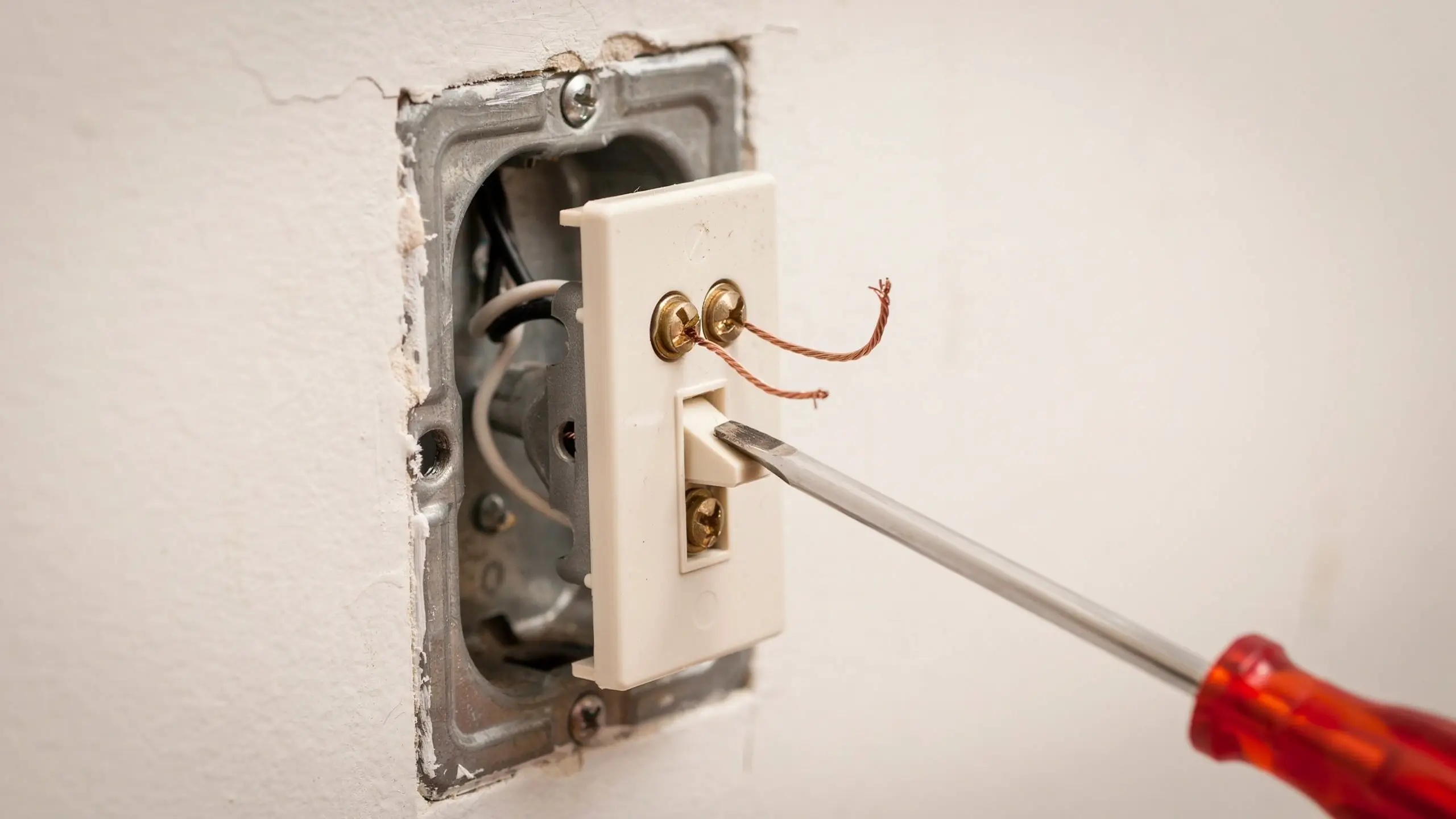 A red-handled screwdriver loosening the bottom screw of a white toggle switch during Step 5: Disconnect the Old Switch, revealing the recessed metal electrical box and copper grounding wires.