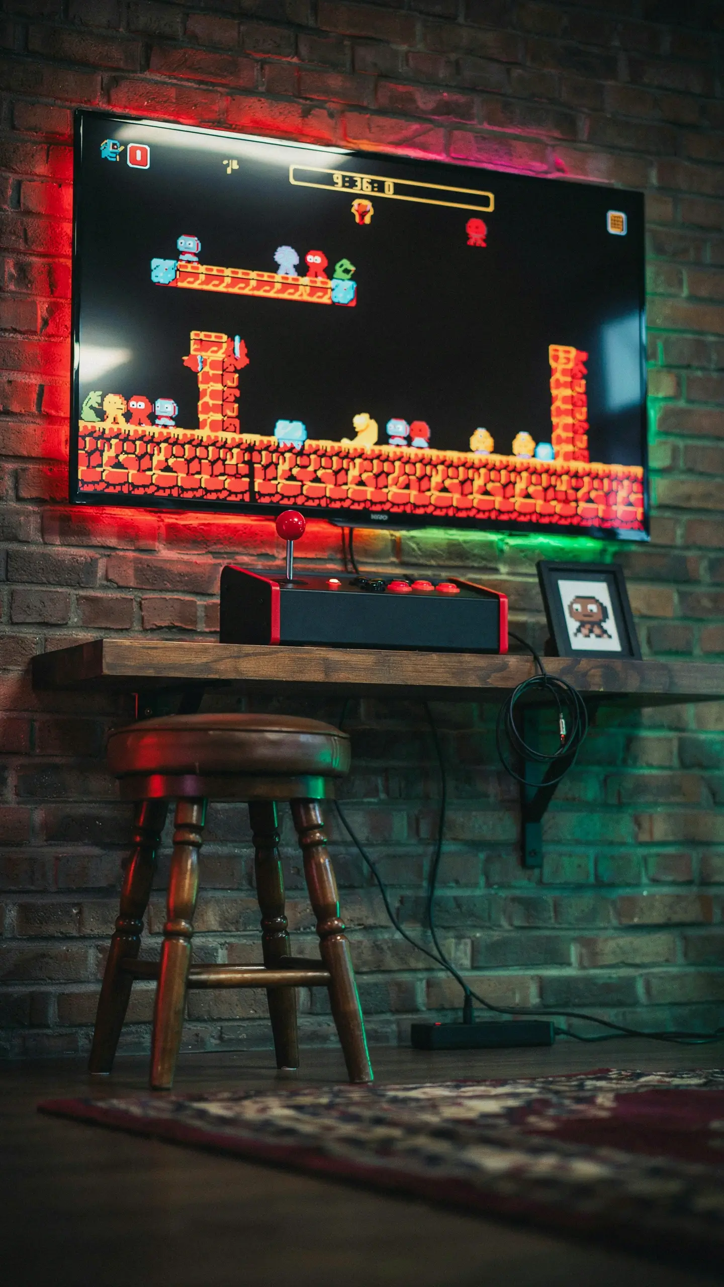 A cozy retro arcade corner with a flat-screen TV mounted on a brick wall, displaying a colorful 8-bit pixel platformer game. The setup features a floating wooden shelf holding a black and red arcade fight stick, paired with a classic wooden stool, framed pixel art, and red and green ambient LED lighting