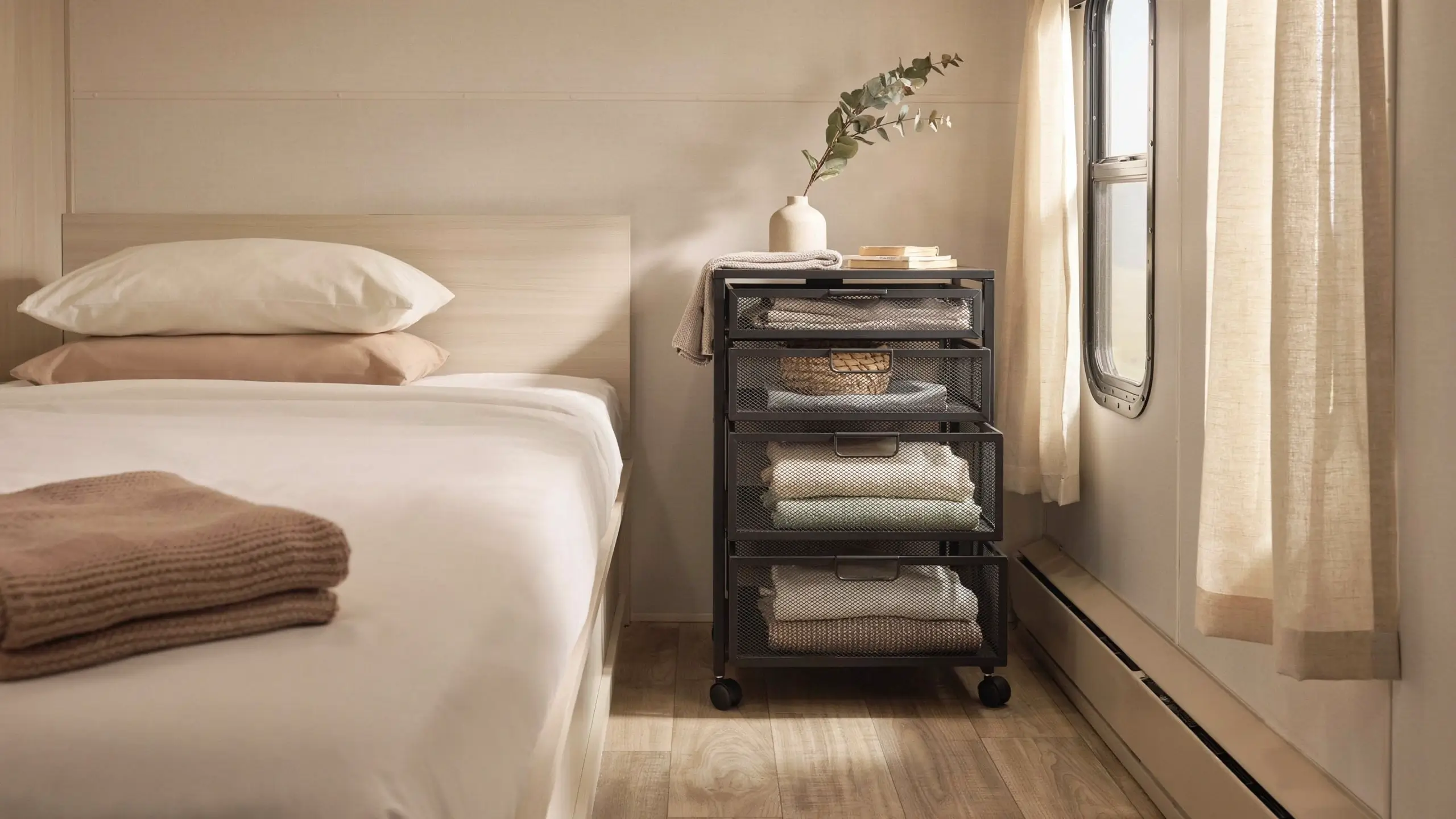 Black rolling mesh drawer cart used as a space-saving nightstand and dresser alternative in a compact RV bedroom with white bedding.