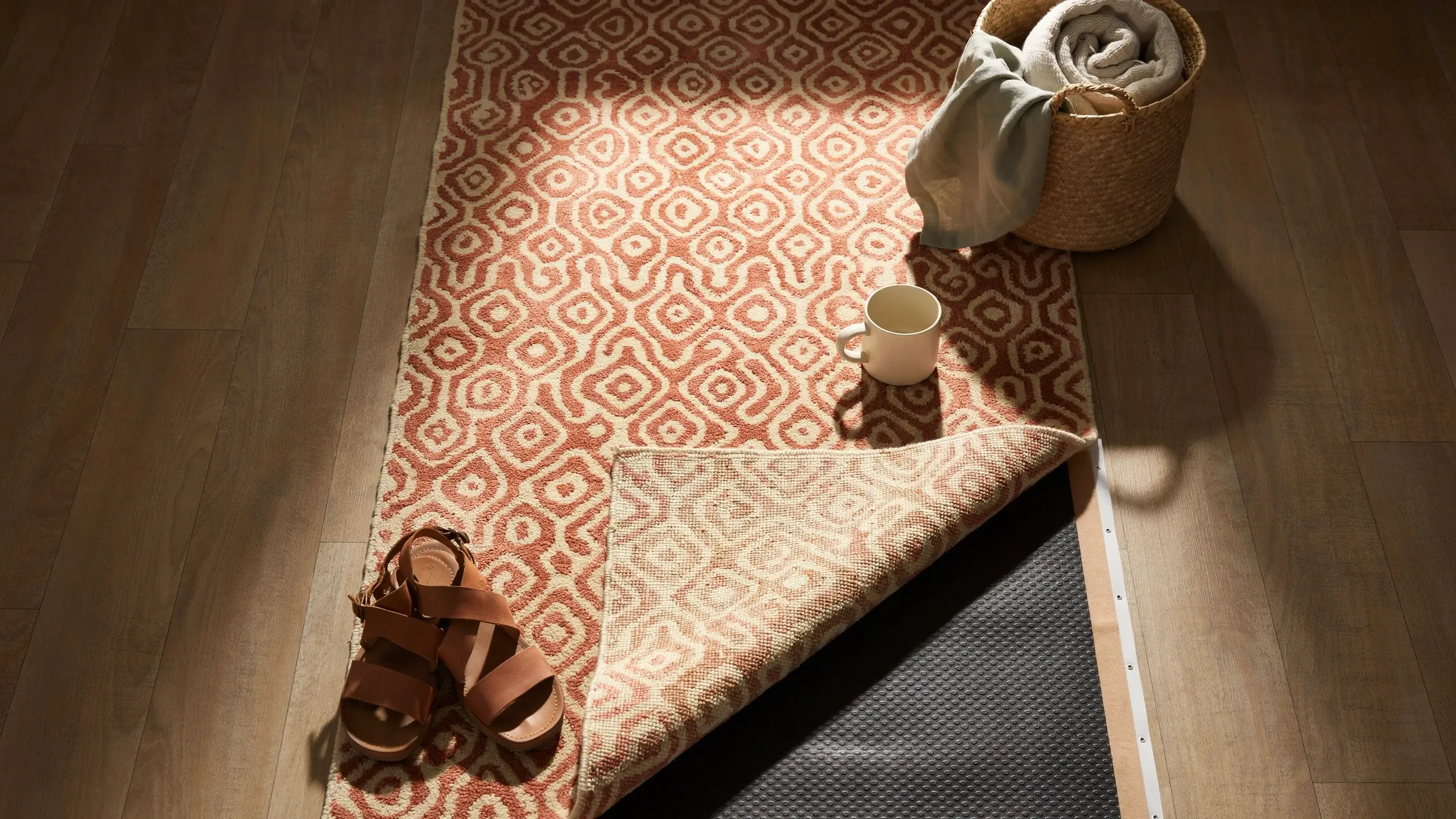 A rust and cream geometric patterned area rug with its corner folded back, revealing a black textured non-slip rug pad underneath to prevent sliding on a light brown hardwood floor, styled with brown leather sandals and a white ceramic mug.