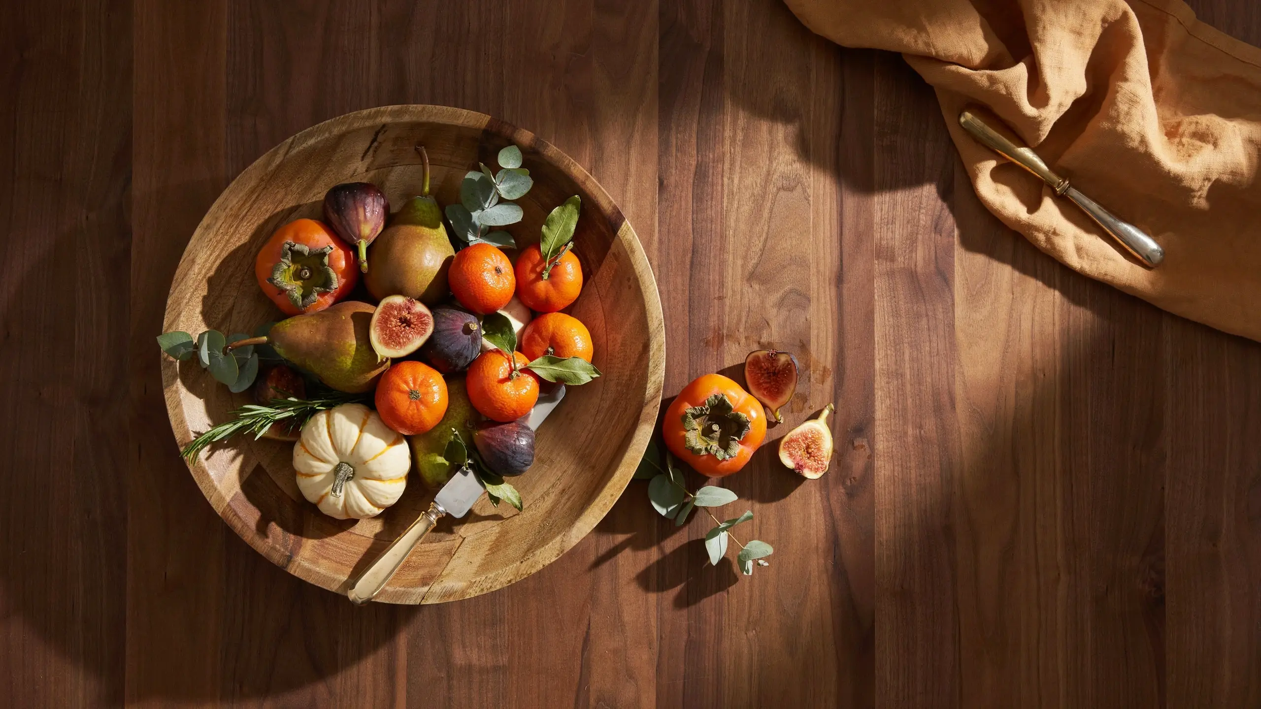 Overhead view of a rustic wooden bowl brimming with autumn fruits like persimmons, sliced figs, and pears, surrounded by a knife and a tan linen cloth on a sunlit wood table.