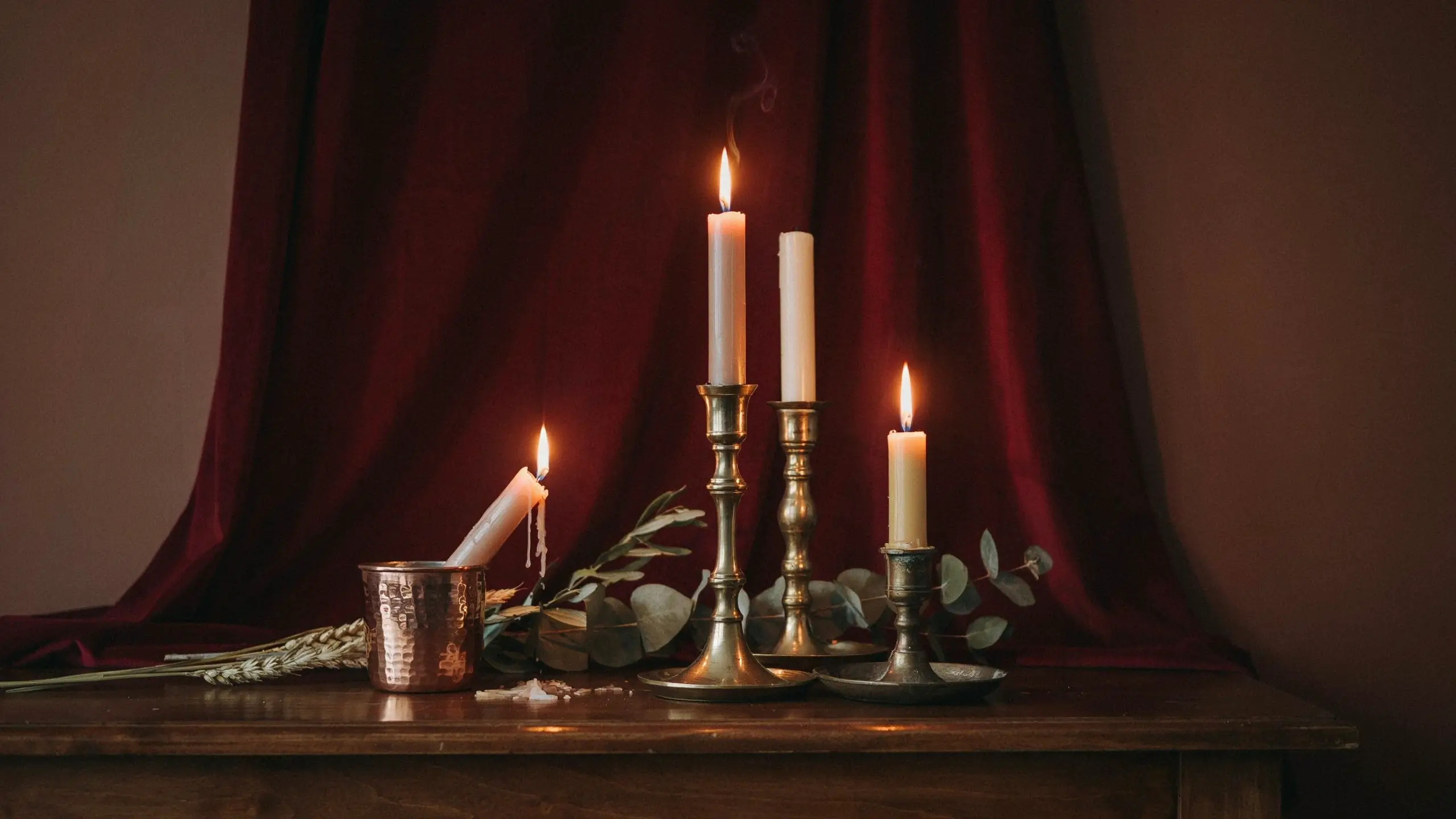 Brass and copper candlesticks with lit white candles, eucalyptus leaves, and dried wheat arranged on a dark wood table against a burgundy velvet curtain.