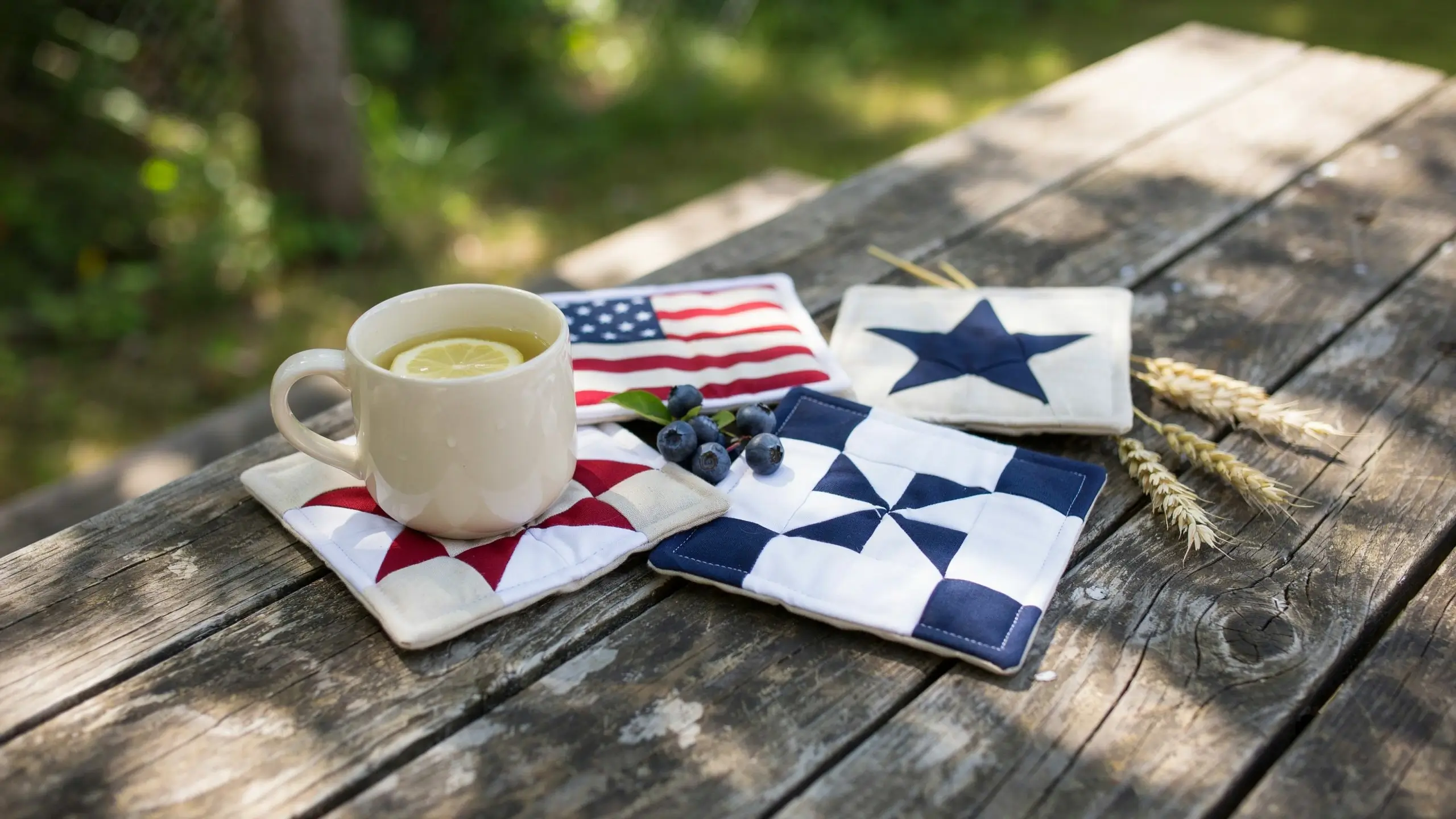 White mug with lemon tea on a handmade red and white flag block mug rug coaster, styled with patriotic fabric coasters, blueberries, and wheat on a rustic wooden table