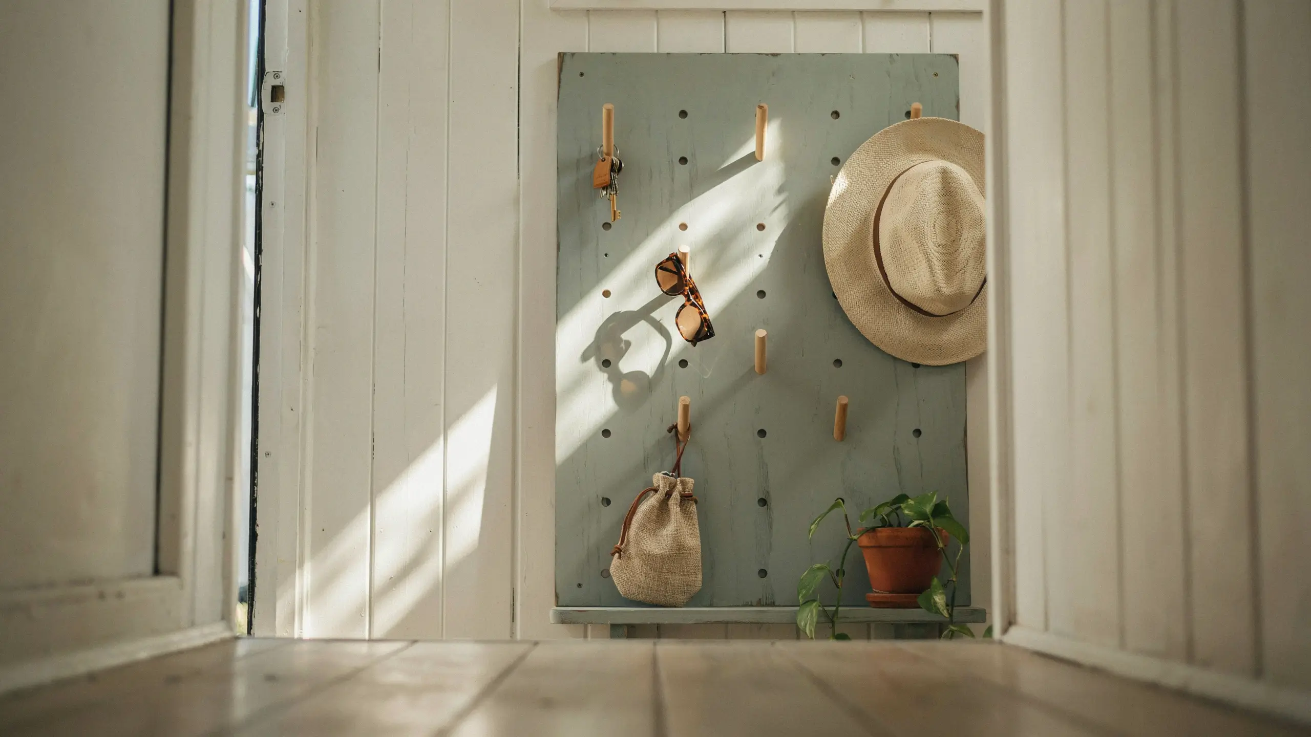 Rustic wooden pegboard organizer in a sunlit white entryway with a hanging straw hat, sunglasses, keys, a small burlap hanging bag for open storage, and a trailing potted plant. 
