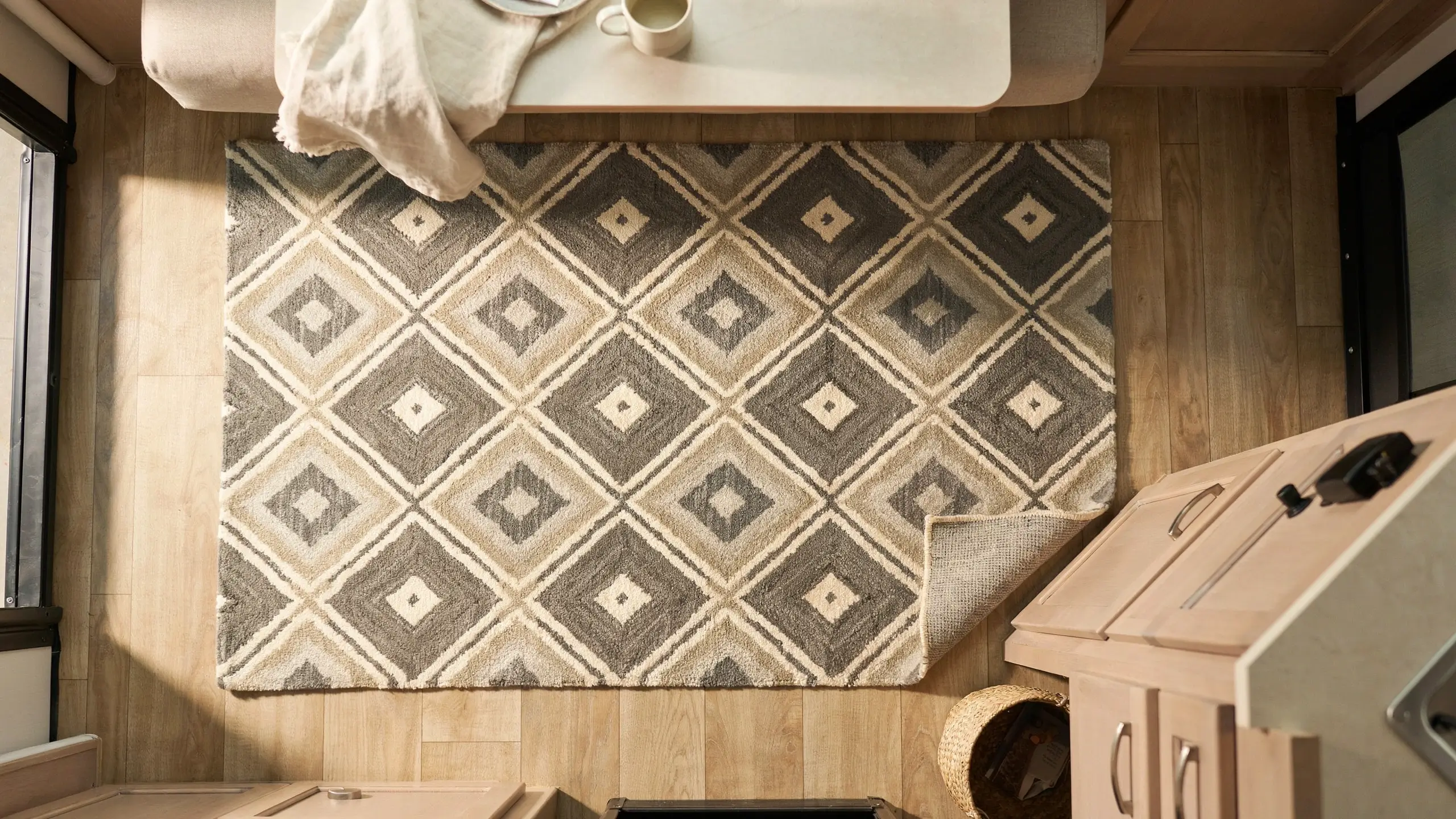 A top-down view of a cozy RV camper dinette area featuring a beige and grey geometric patterned area rug on light wood flooring. The white dinette table holds a mug and cream-colored fabric, providing an inspiration shot for pairing a new patterned rug with newly reupholstered or slipcovered dinette cushions during a motorhome renovation. 