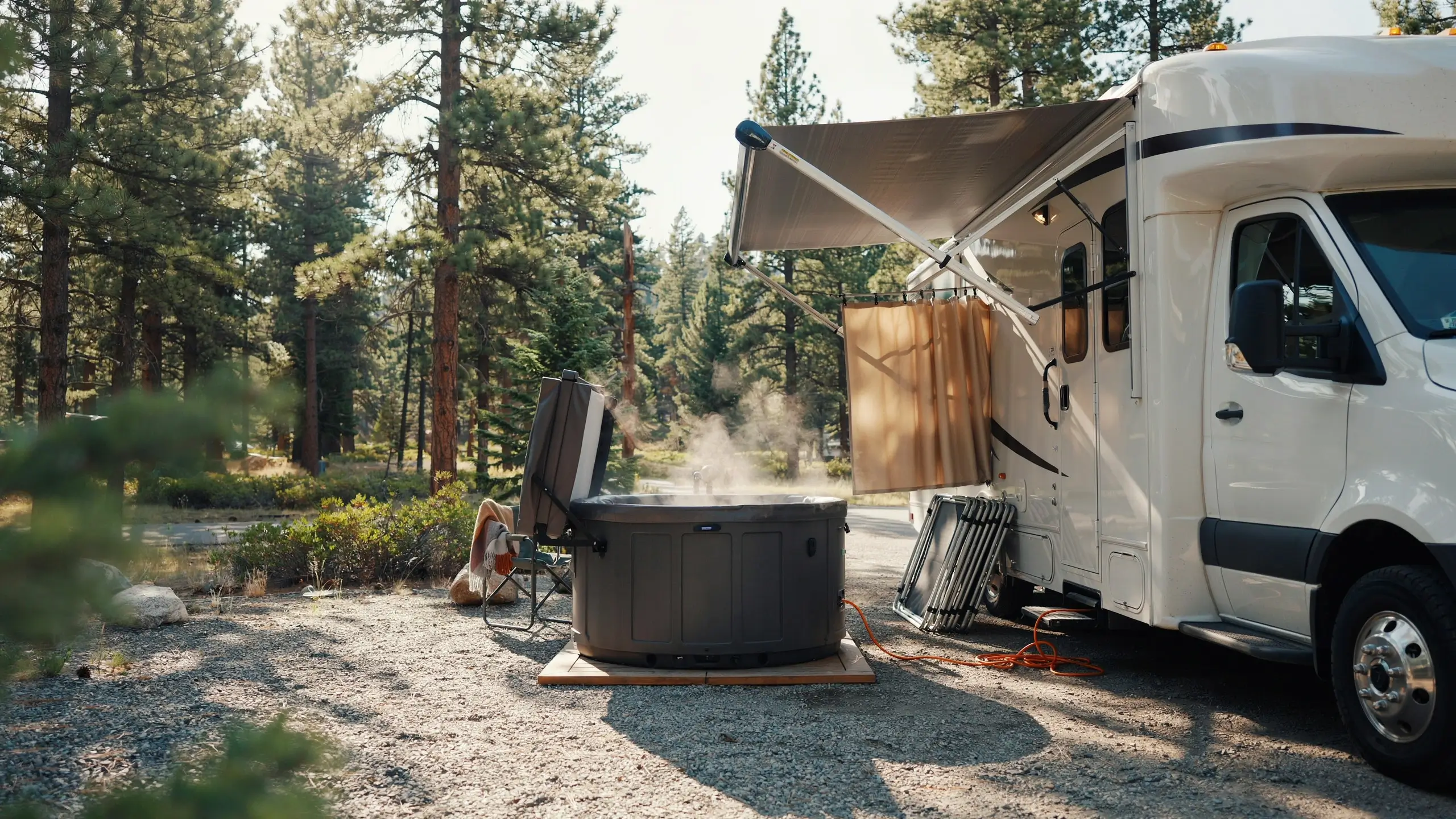 White RV camper with a simple privacy curtain hanging from the awning, creating a secluded space next to a steaming portable hot tub resting on a wooden base in a pine forest campsite.