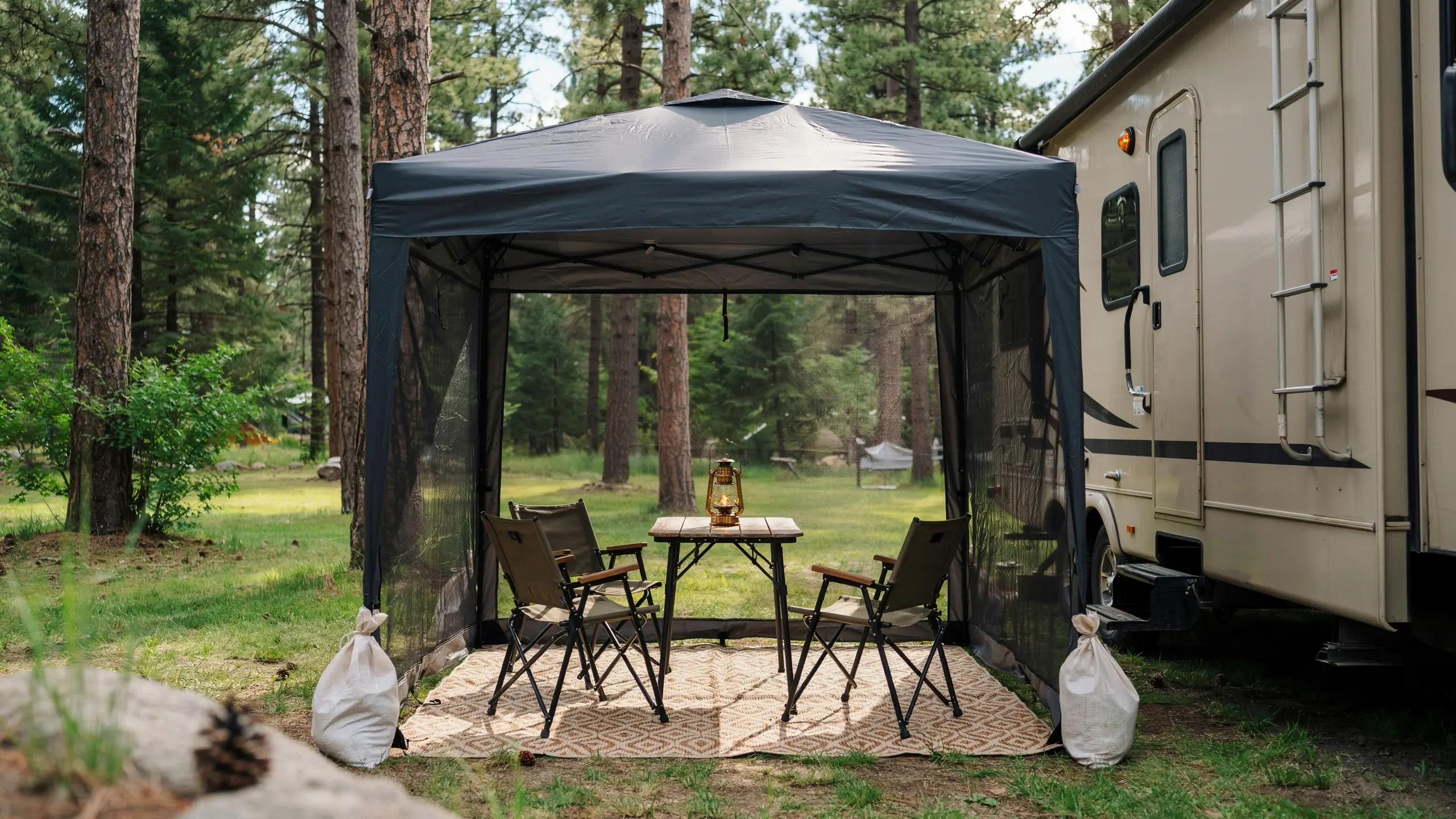 Dark grey popup screen tent with chairs and a lantern, set up on a rug next to an RV in a pine forest for a bug-free outdoor room. 