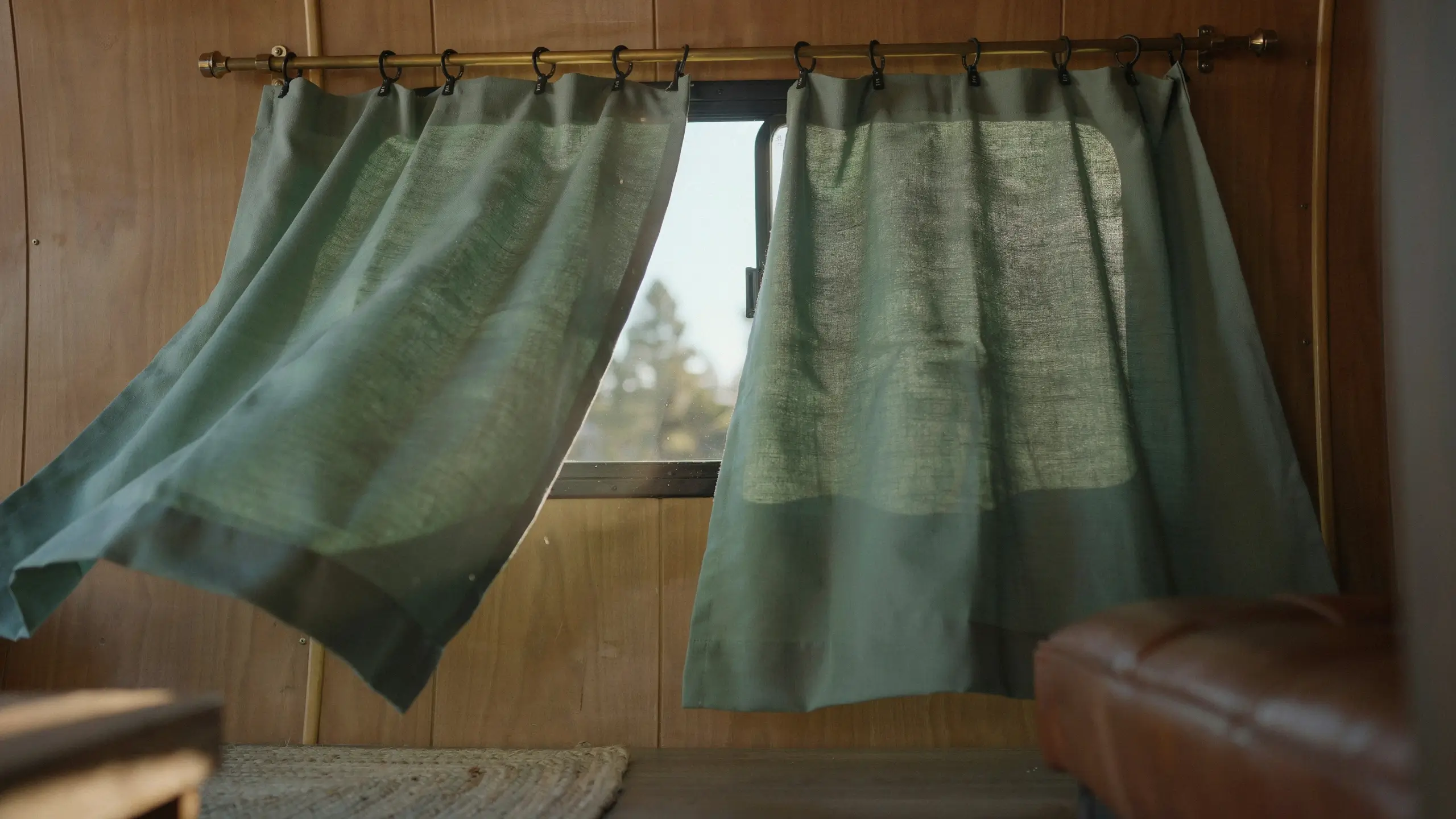Sage green clip-on curtains hanging on a brass rod inside a wooden van interior, parted to reveal a window with pine trees, being tested before a road trip.