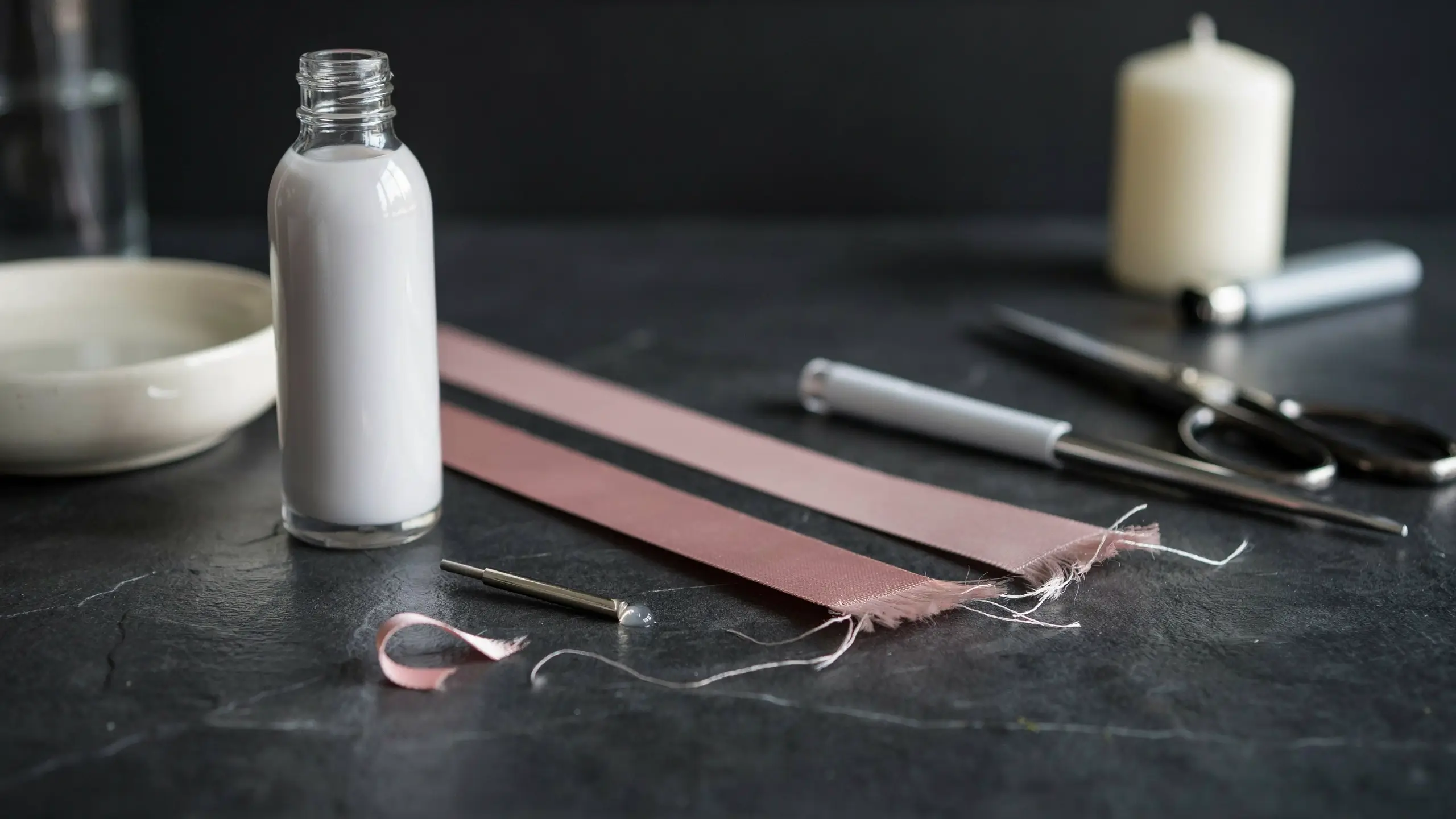 Alt Text: Pink ribbon strips with frayed ends beside scissors, fabric sealant, a candle, and a lighter for edge sealing.