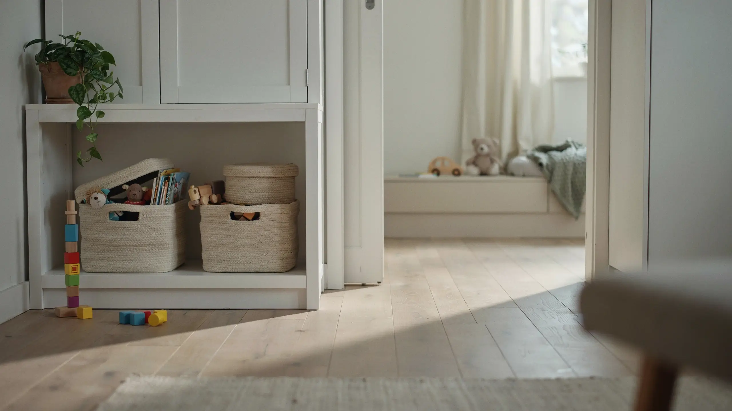 Minimalist white built-in storage holding woven baskets for toy rotation, wooden building blocks on floor, and potted plant in sunlit playroom. 