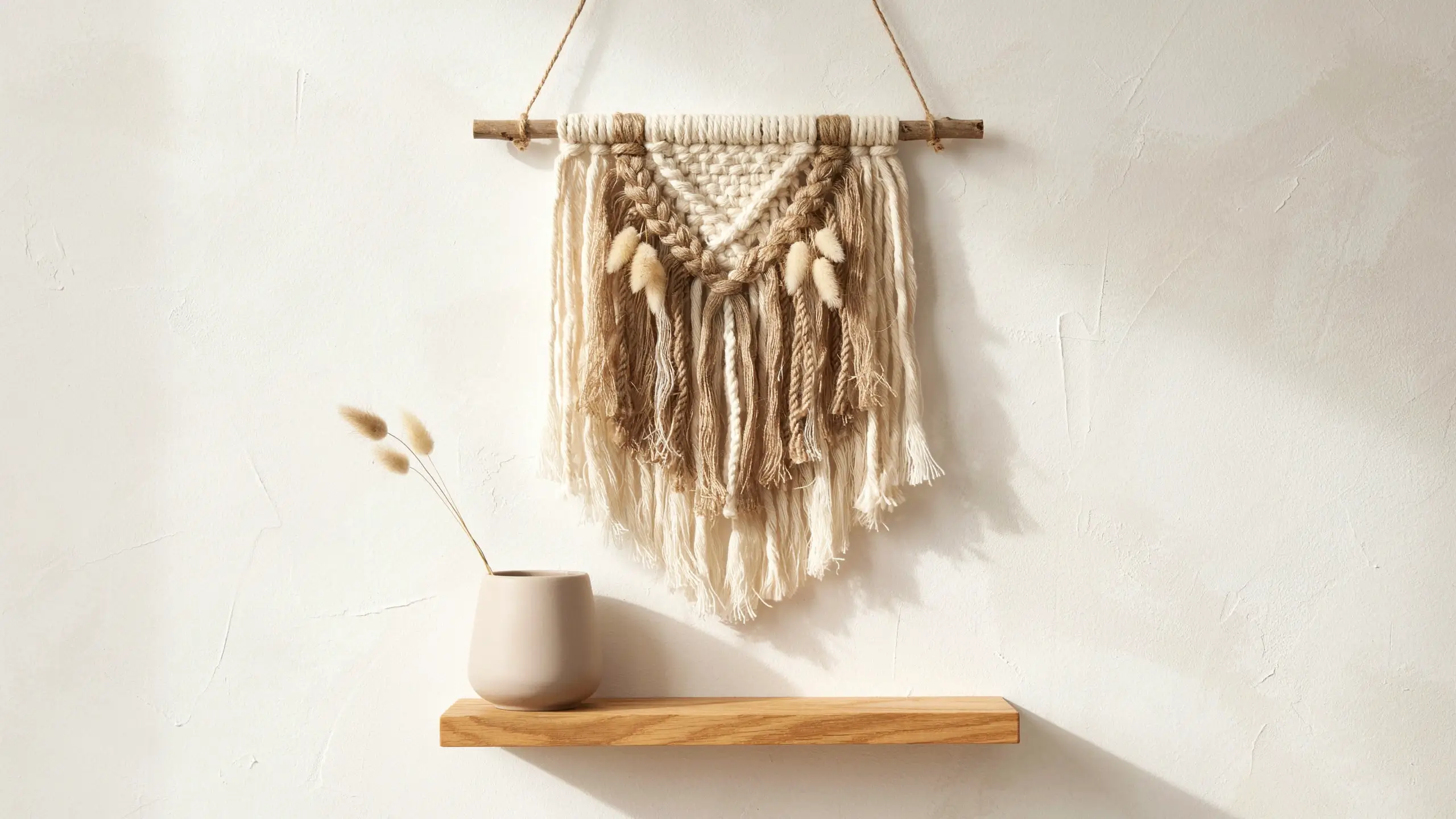 Minimalist boho macrame yarn wall hanging with wooden dowel and dried bunny tail accents displayed over a small floating shelf with a beige vase.