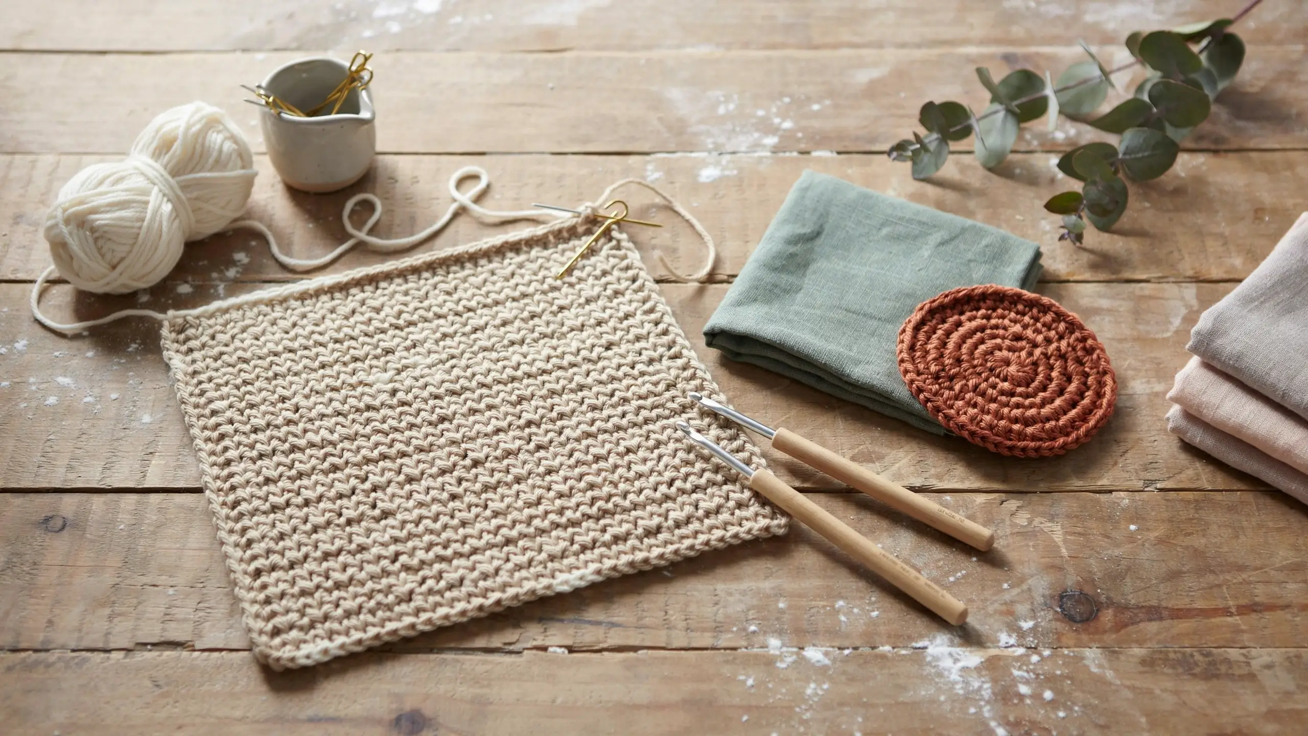 A beige single crochet stitch (sc) swatch resting on a rustic wooden table, arranged with two wooden-handled crochet hooks, a white yarn ball, gold stitch markers, a sage green folded linen napkin, a rust-colored round crochet coaster, and a sprig of eucalyptus in a cozy flat-lay craft setting.