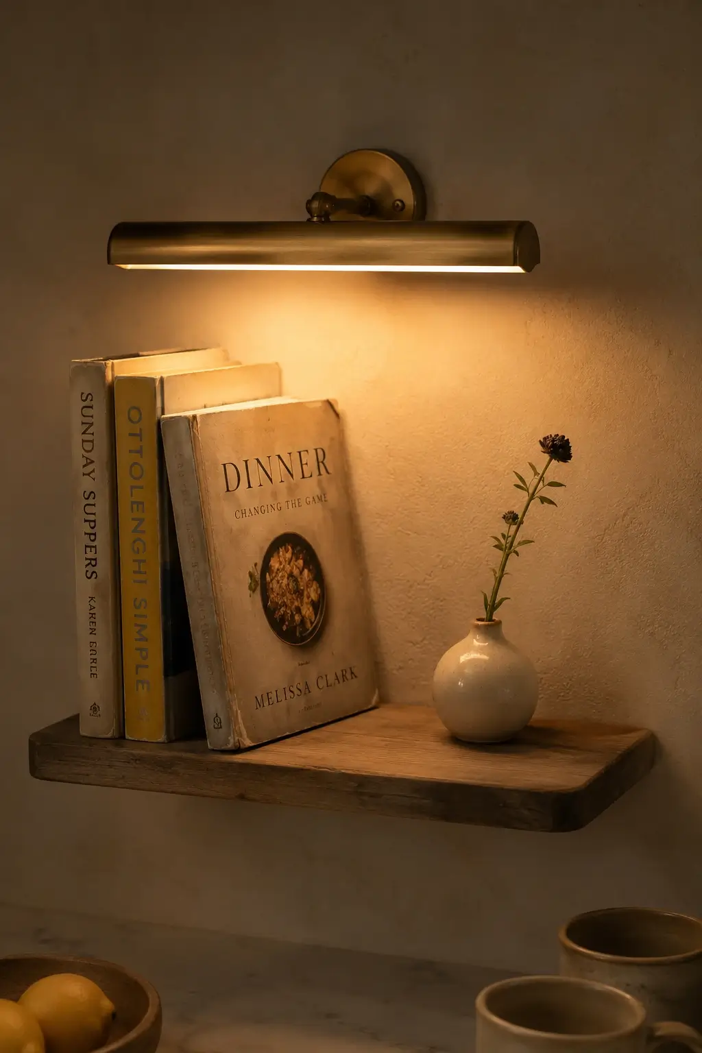 Open cookbook shelf with a slim brass picture light, warm glow, worn cookbooks, and a small ceramic bud vase.