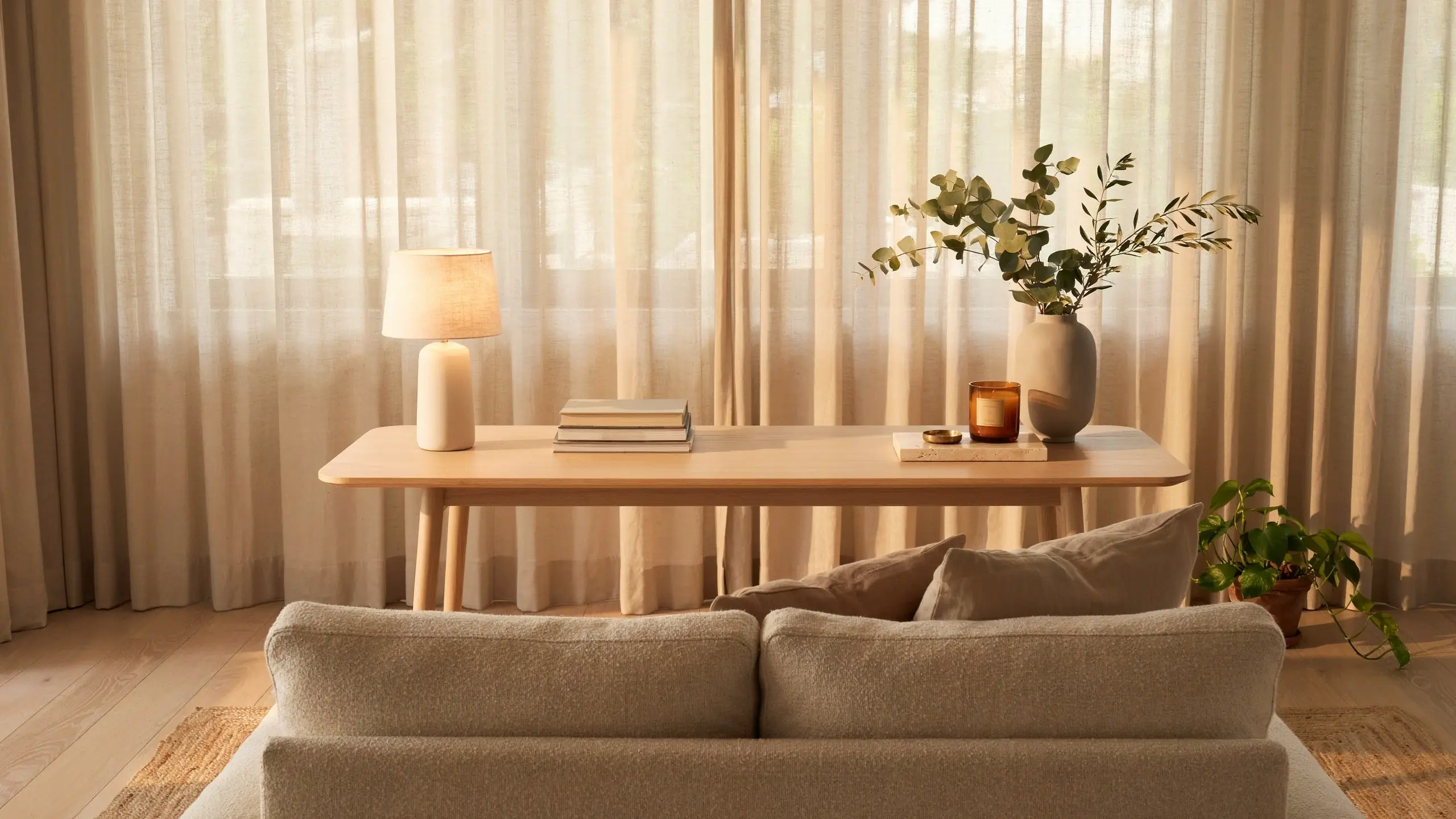 Minimalist light wood slim sofa table placed behind a textured beige couch, styled with a ceramic lamp, books, amber candle, and a tall vase of eucalyptus. 