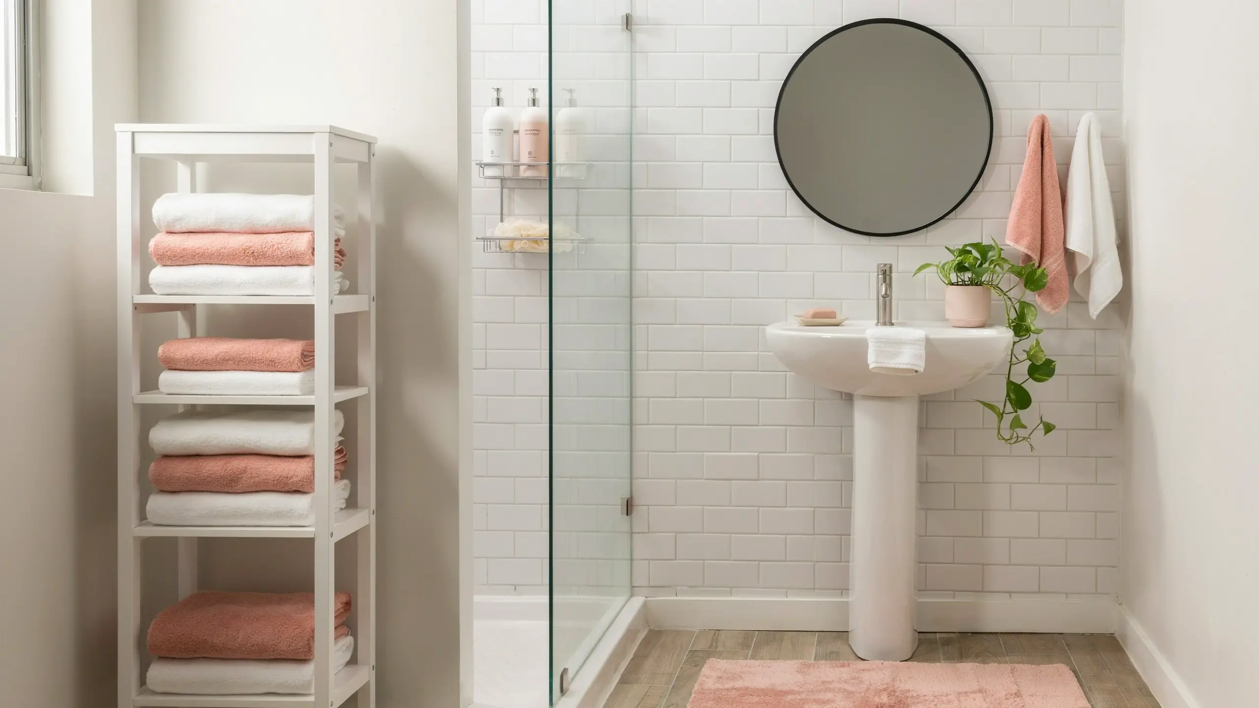 Bright small bathroom design ideas with white subway tile walls, a glass-enclosed shower, and a white pedestal sink paired with a large round mirror. An open white shelving unit neatly holds folded pink and white towels, with a potted pothos plant adding greenery to the space