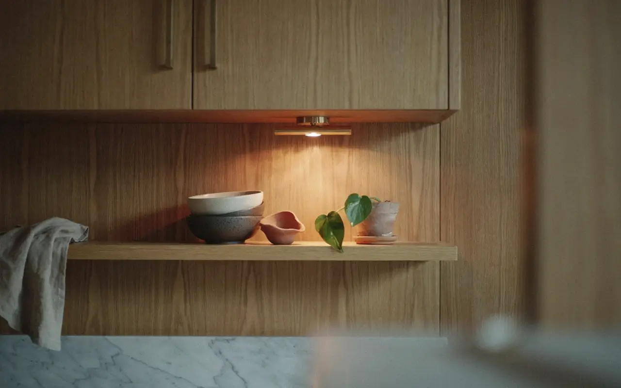 Small picture light above an open wood kitchen shelf, giving bowls and a plant a warm gallery-lit glow.