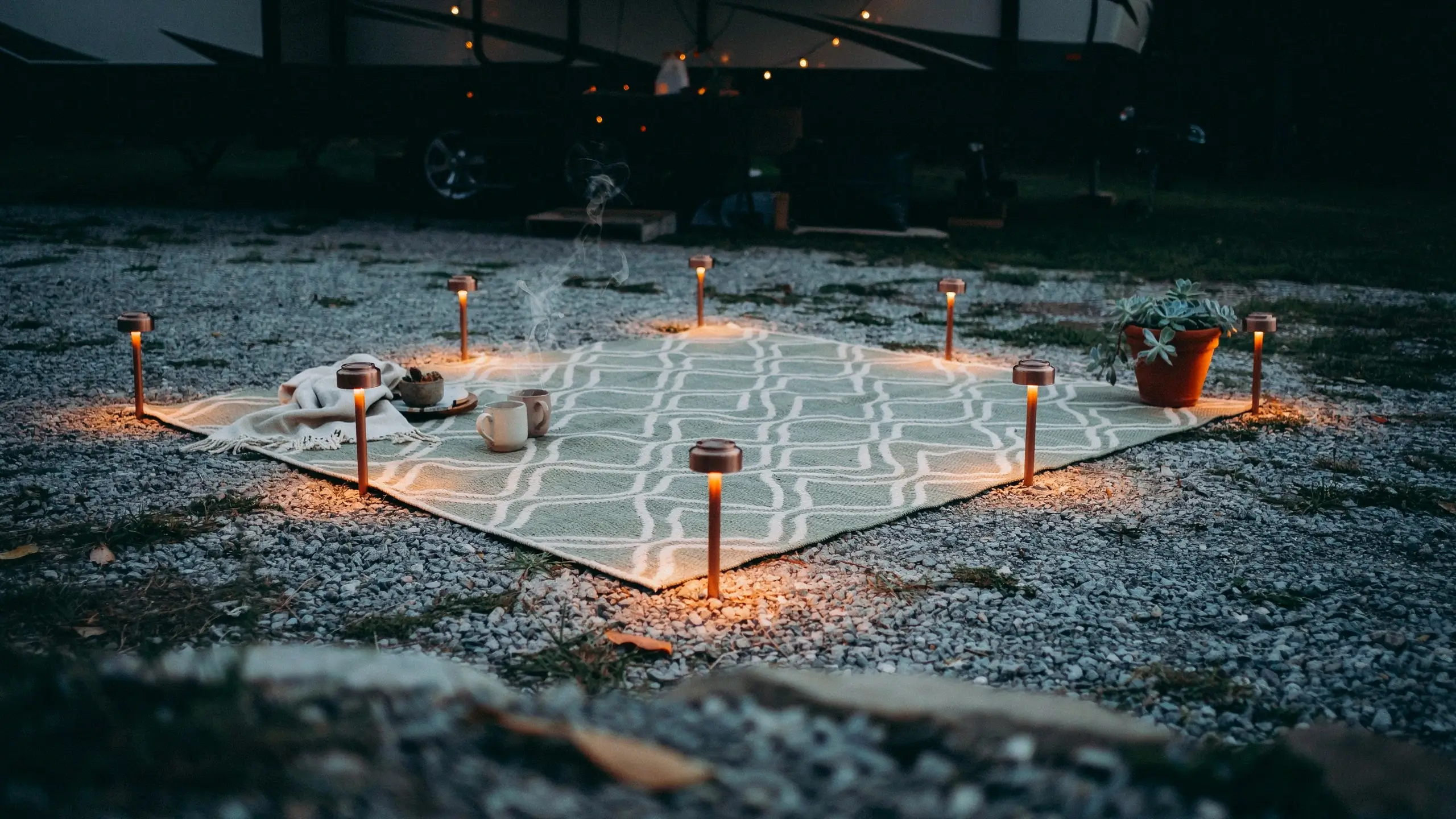 "Cozy RV campsite setup at dusk featuring warm solar path lights arranged around the perimeter of a geometric outdoor rug on a gravel ground." 