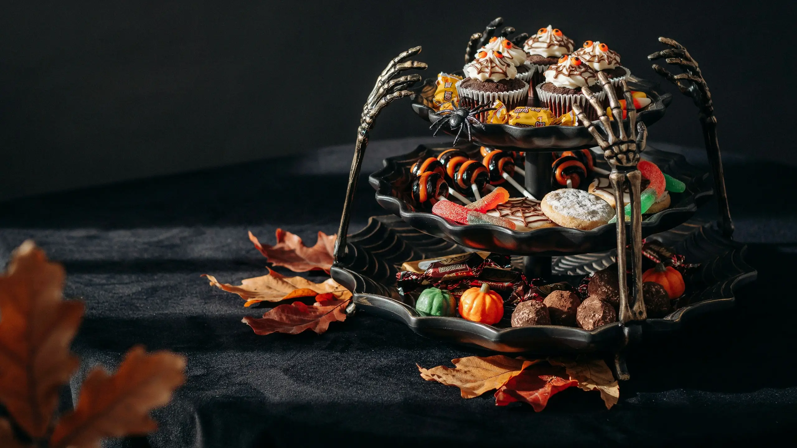 A three-tiered Halloween dessert stand with skeleton arm supports holding spooky cupcakes with candy eyes, swirled lollipops, candy corn, gummy sweets, and chocolate truffles, displayed on a dark black cloth surrounded by autumn leaves. 