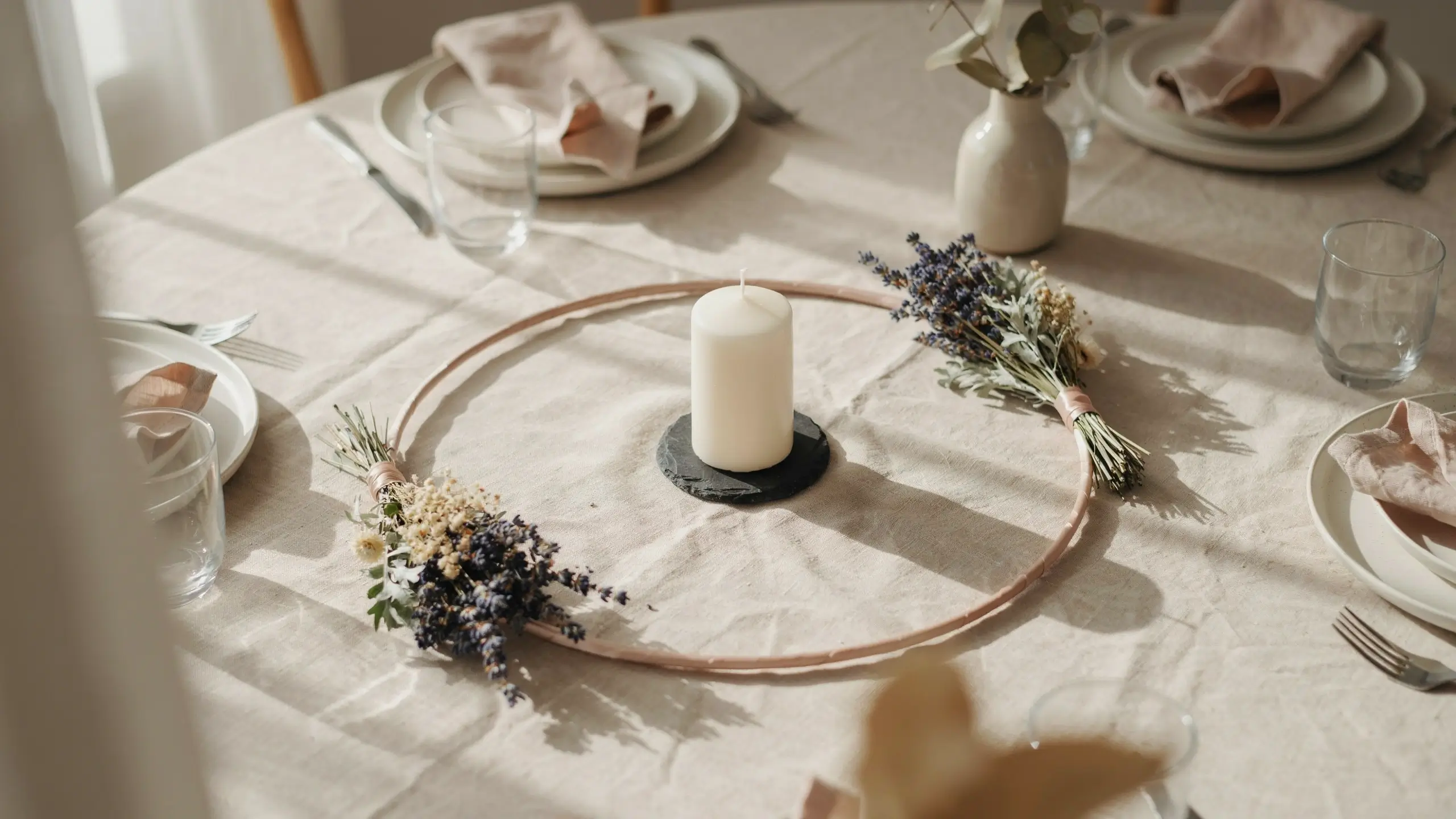 A minimalist spring table centerpiece featuring a wooden floral hoop decorated with dried lavender and white filler flowers, surrounding a white pillar candle on a slate coaster, set on a beige linen tablecloth. 