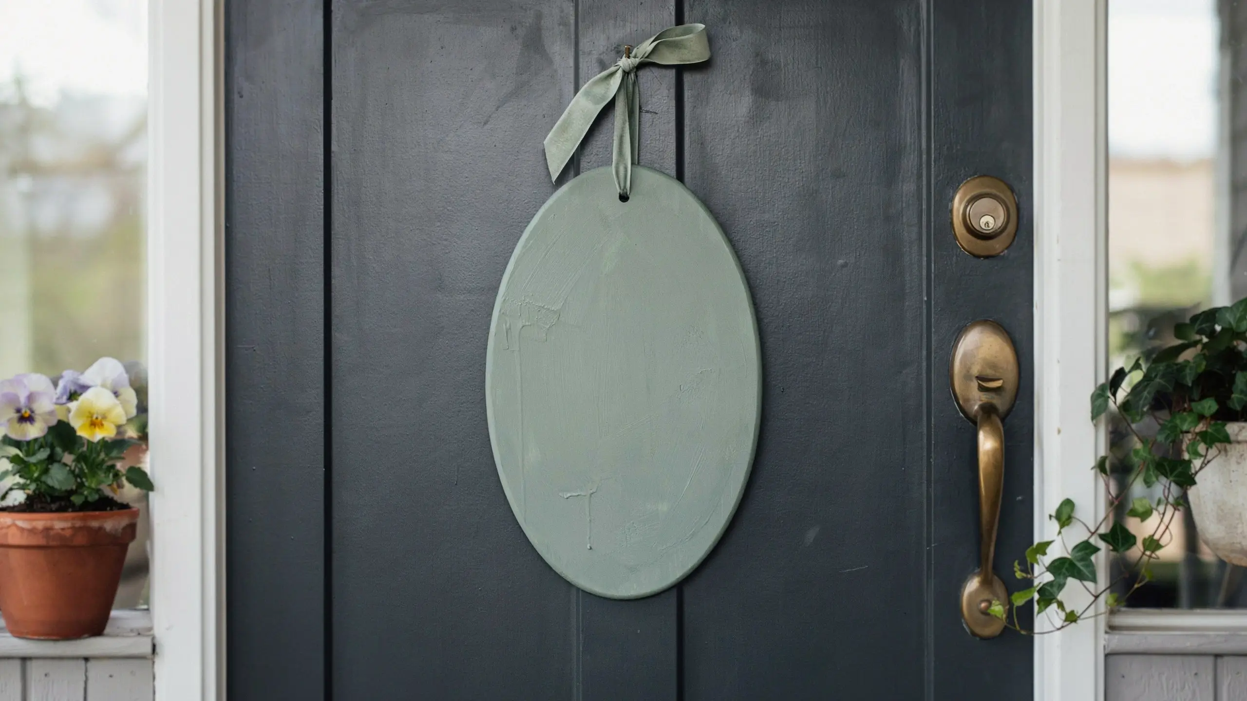 A blank, sage green oval wooden sign hanging from a dark plank front door with a matching velvet ribbon, situated next to an antique brass handle and terracotta potted pansies for a spring porch display. 