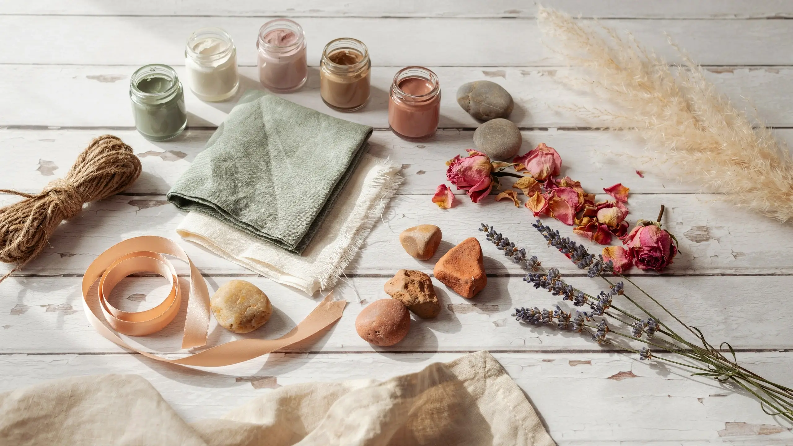 Overhead flat lay of small jars with pastel green, pink, and brown natural clay pigments, styled with dried lavender, pink roses, pampas grass, sage green linen napkins, and jute twine on a rustic white wooden table. 