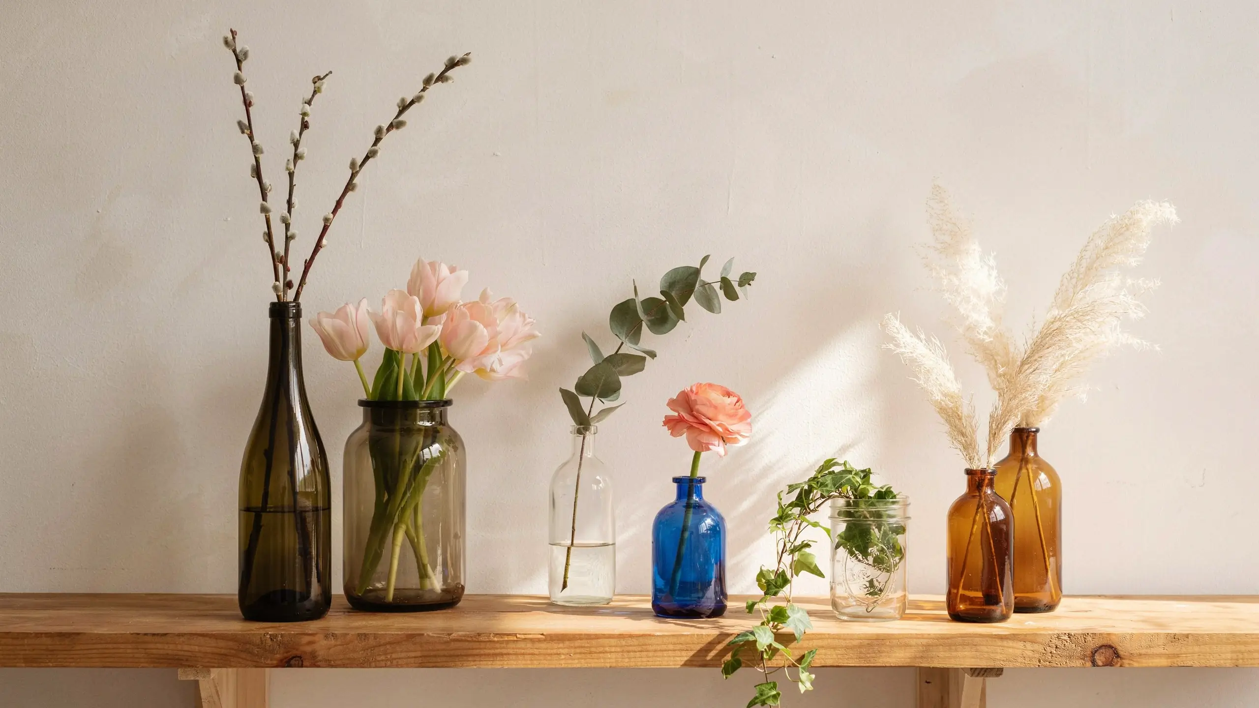 A sunlit spring vignette featuring repurposed thrift store glass bottles holding grocery store floral stems, including pink tulips, pussy willow, eucalyptus, and pampas grass displayed on a rustic wooden shelf.