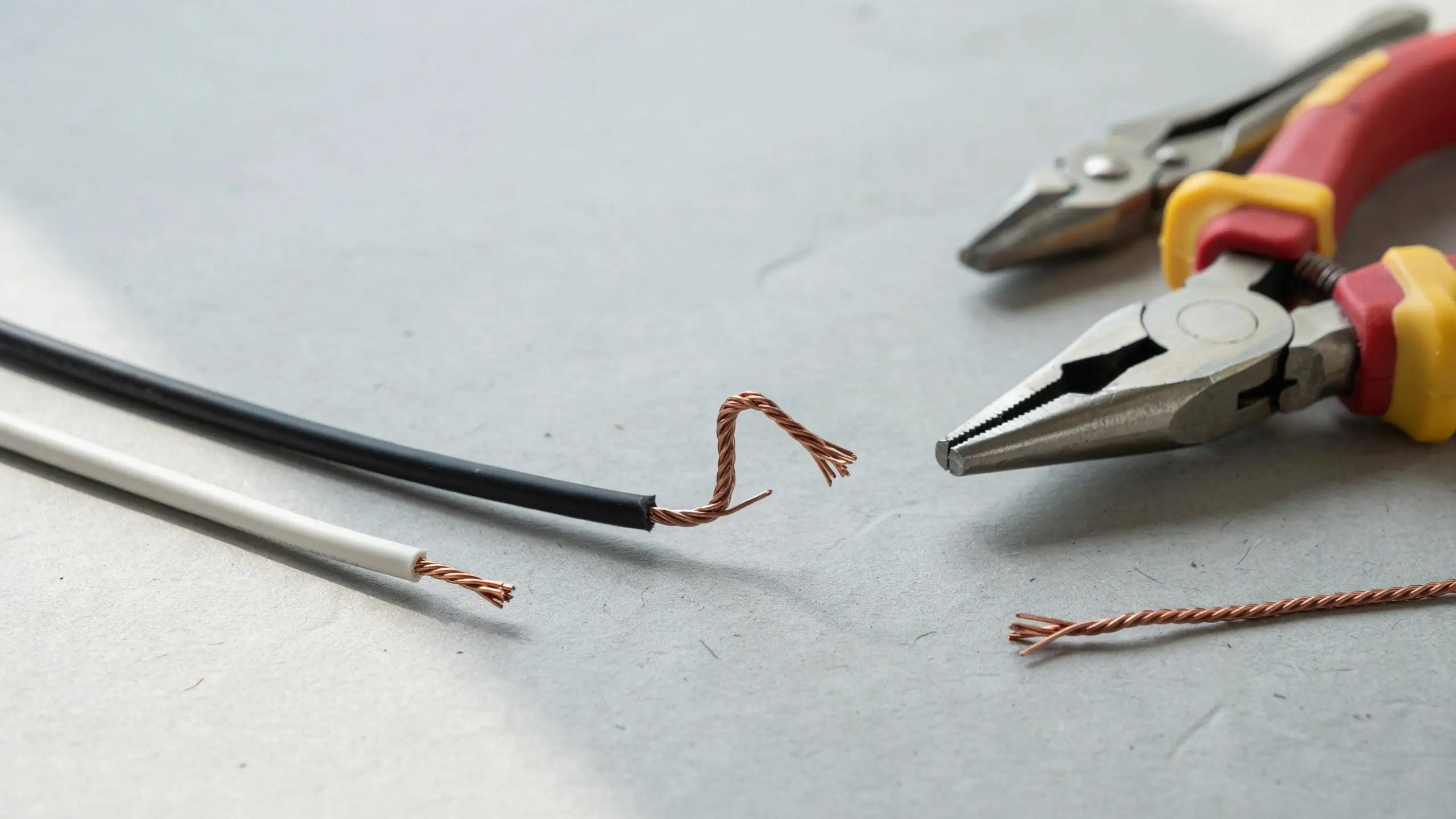 Close-up of white and black electrical wires with stripped insulation exposing twisted copper cores, positioned beside red-handled needle-nose pliers during a DIY wiring preparation step.