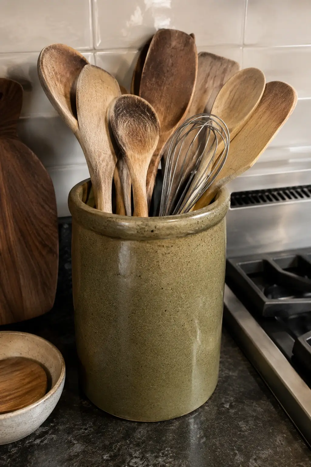 Wooden spoons and whisk in an olive stoneware crock beside a kitchen stove.