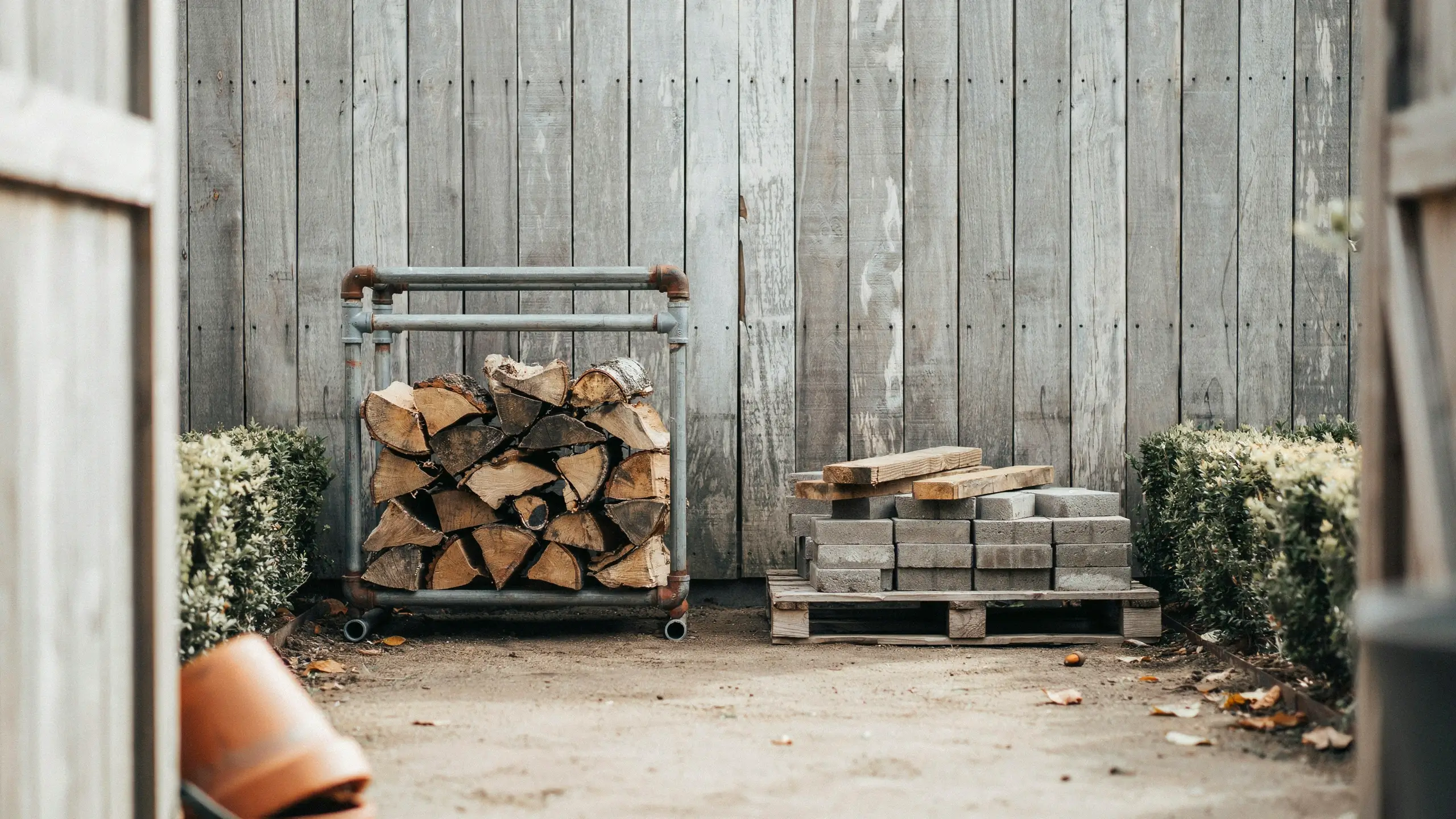 Rustic backyard scene showing a metal pipe rack storing chopped firewood off the ground on a dirt surface. Next to it, gray concrete blocks and wooden planks are neatly stacked on a wooden pallet to keep them elevated and dry.