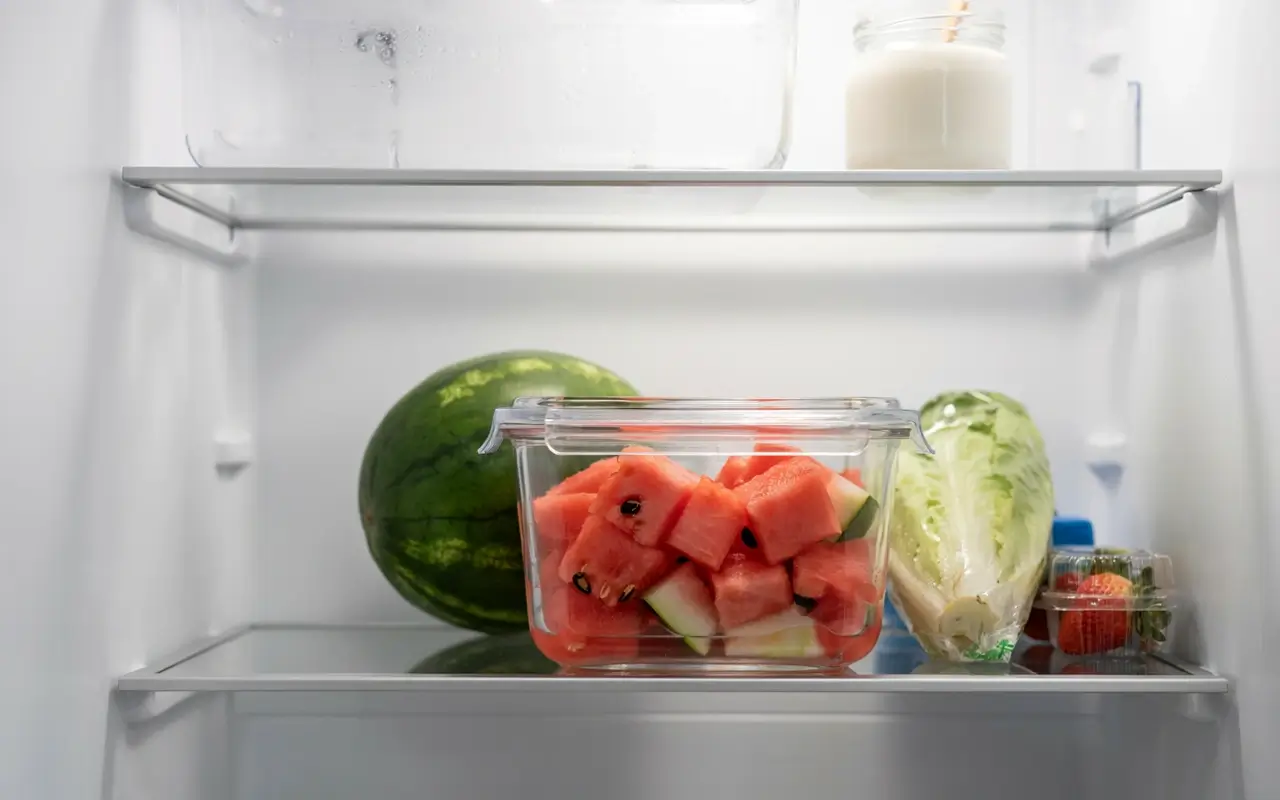Cut watermelon stored in an airtight container inside a refrigerator with a whole melon and fresh produce