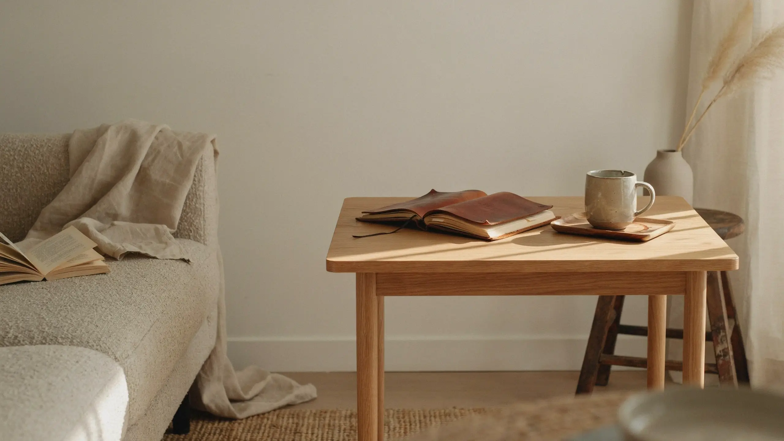 Sunlit minimalist living room featuring a beige boucle sofa with an open book, a light wood coffee table holding an open leather journal and a ceramic mug, and pampas grass decor.