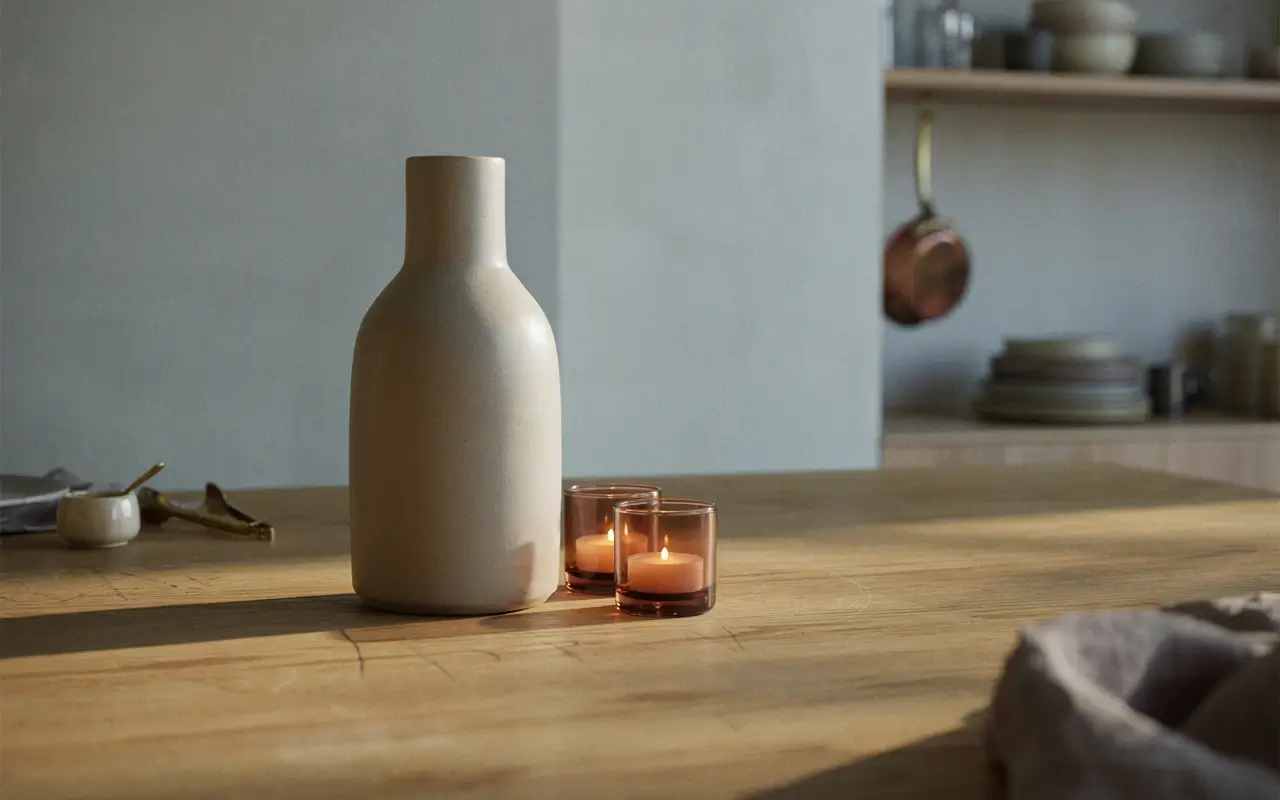 Tall ceramic vase with two glowing amber votives on a wood kitchen table for rule-of-three balance.
