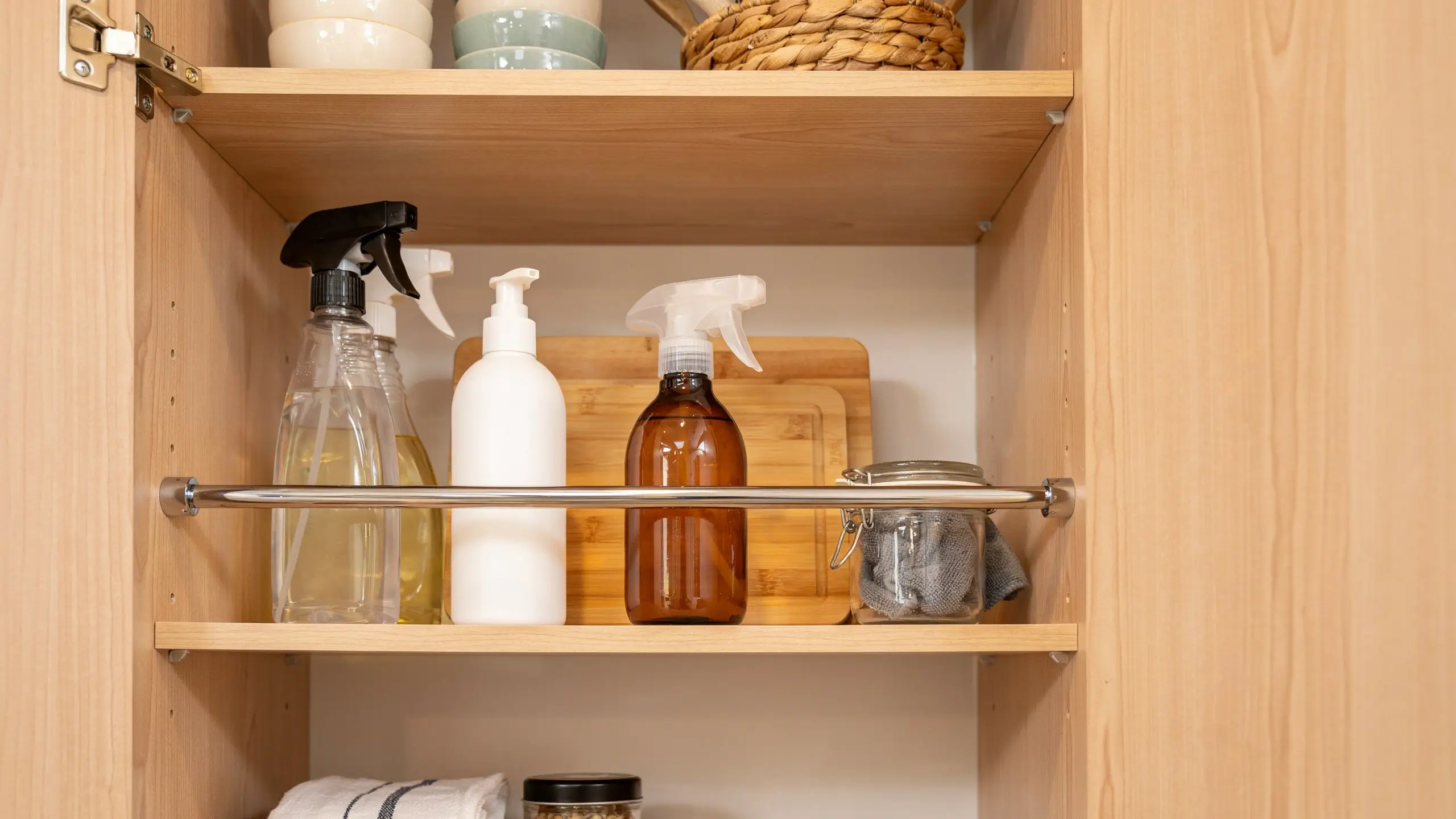 Light wood kitchen cabinet shelf featuring a metal tension rod installed as a protective guardrail to keep cleaning supply bottles and an amber spray bottle from sliding forward and falling