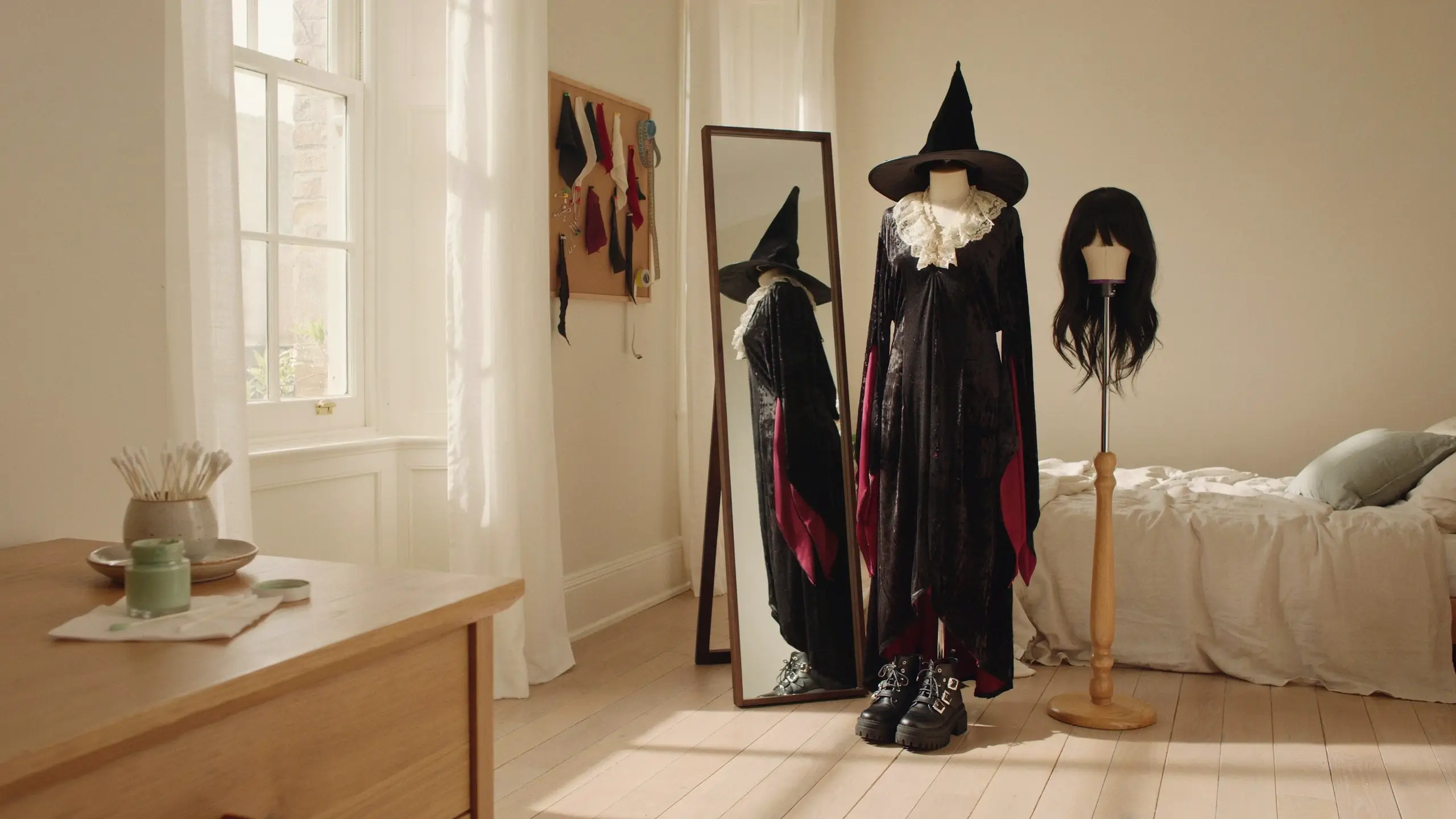 Black velvet witch costume with a white ruffled lace collar and classic pointed hat displayed on a mannequin in a brightly lit bedroom, paired with a matching wig on a wooden stand and chunky black platform boots. 