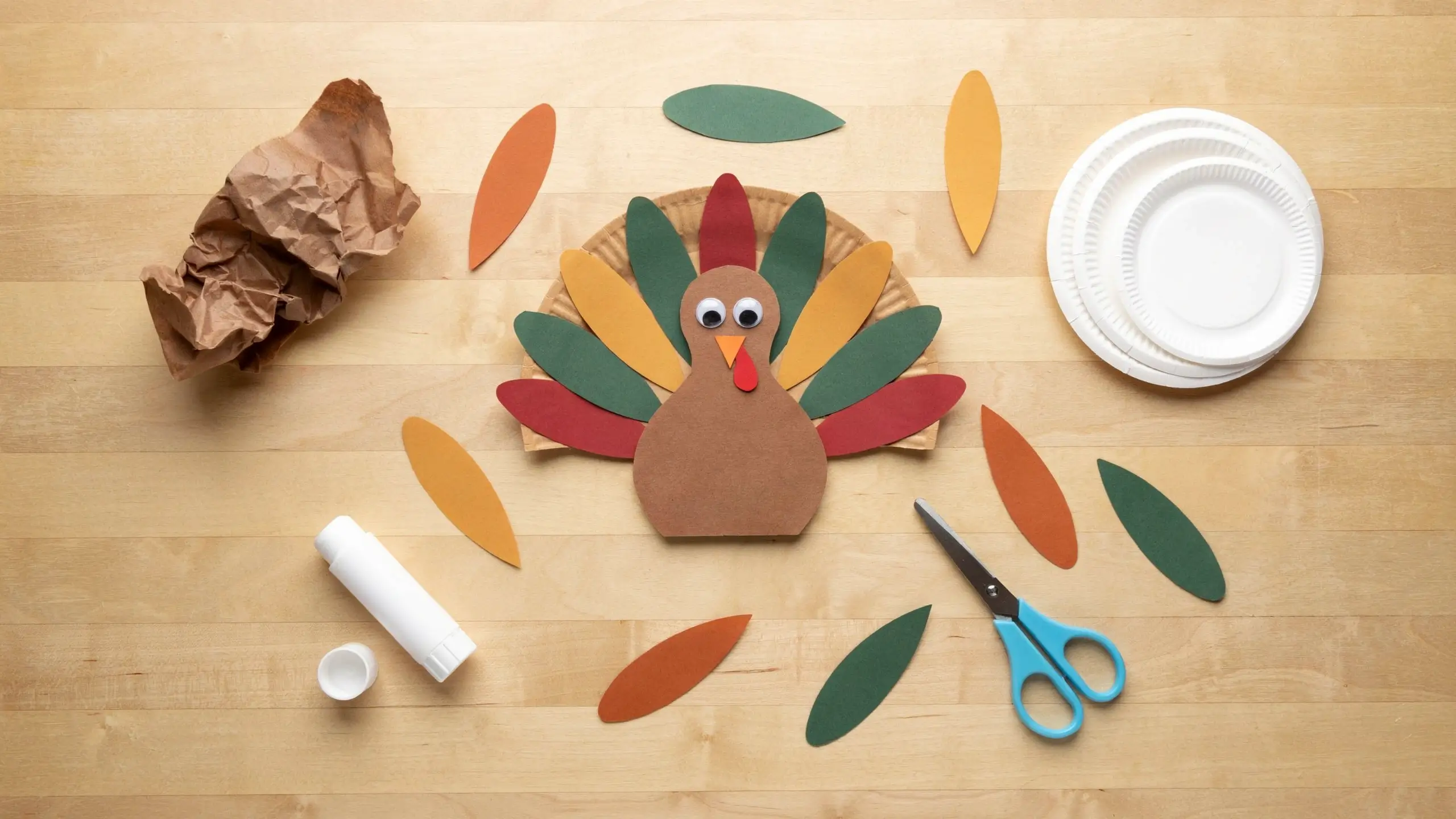 Top-down view of a DIY paper plate turkey craft with colorful paper feathers, googly eyes, and craft supplies like a glue stick and scissors on a wooden table for a fall preschool activity