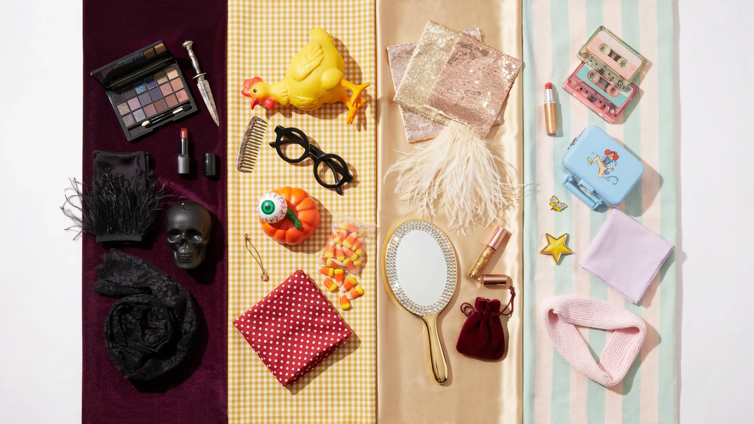 "A vibrant aesthetic flat lay displaying a diverse range of costume accessories, makeup palettes, and props organized on a colorful striped fabric. The visual segmentation features a spooky gothic skull and dark eyeshadow alongside a rustic yellow chicken and candy corn, contrasted with sparkling glamour sequins and rhinestones, and finally a retro pastel section with cassette tapes and a vintage tin lunchbox." 