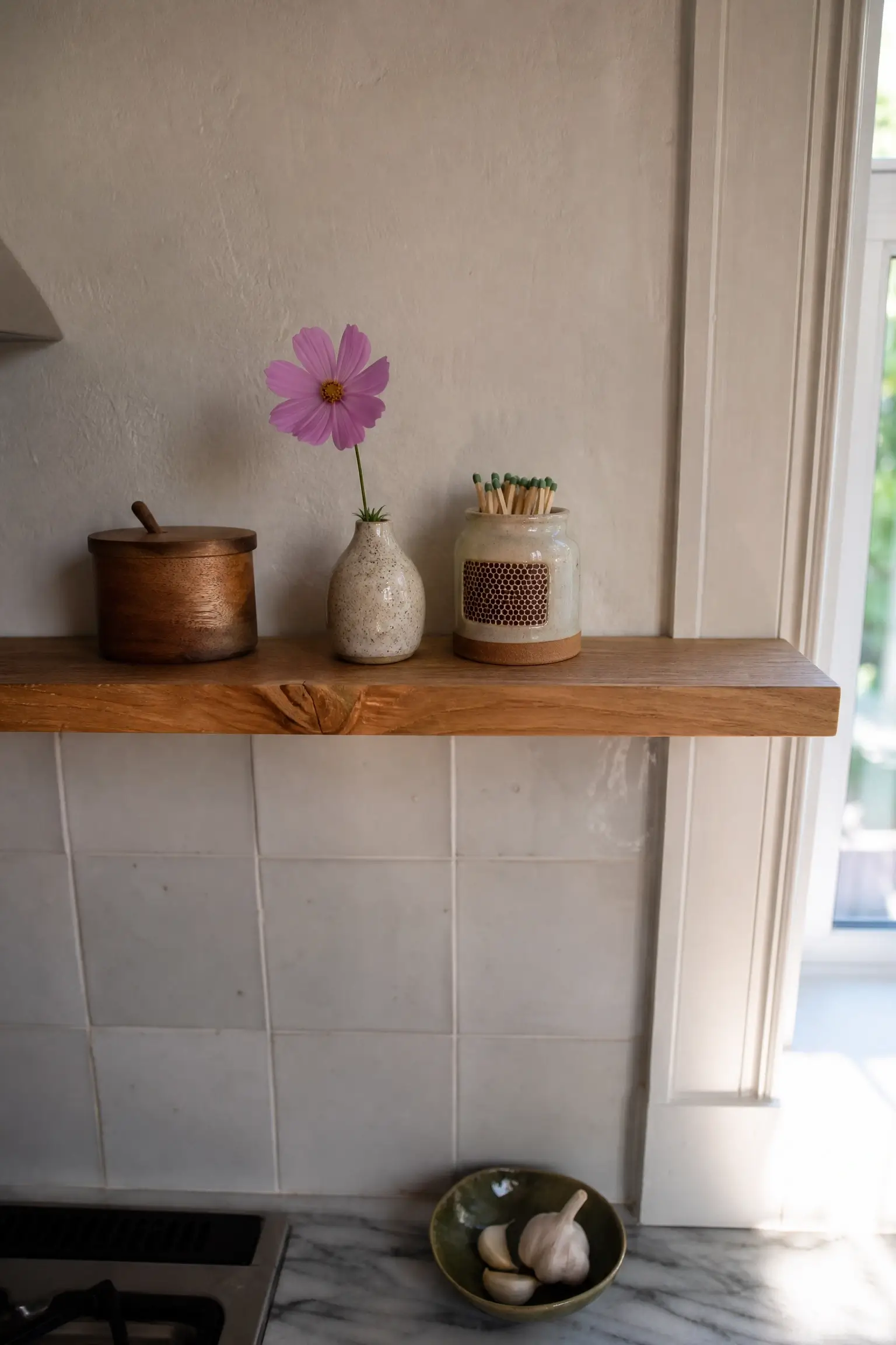 Tiny floating wood shelf above kitchen backsplash with salt cellar, flower vase, and ceramic match striker.