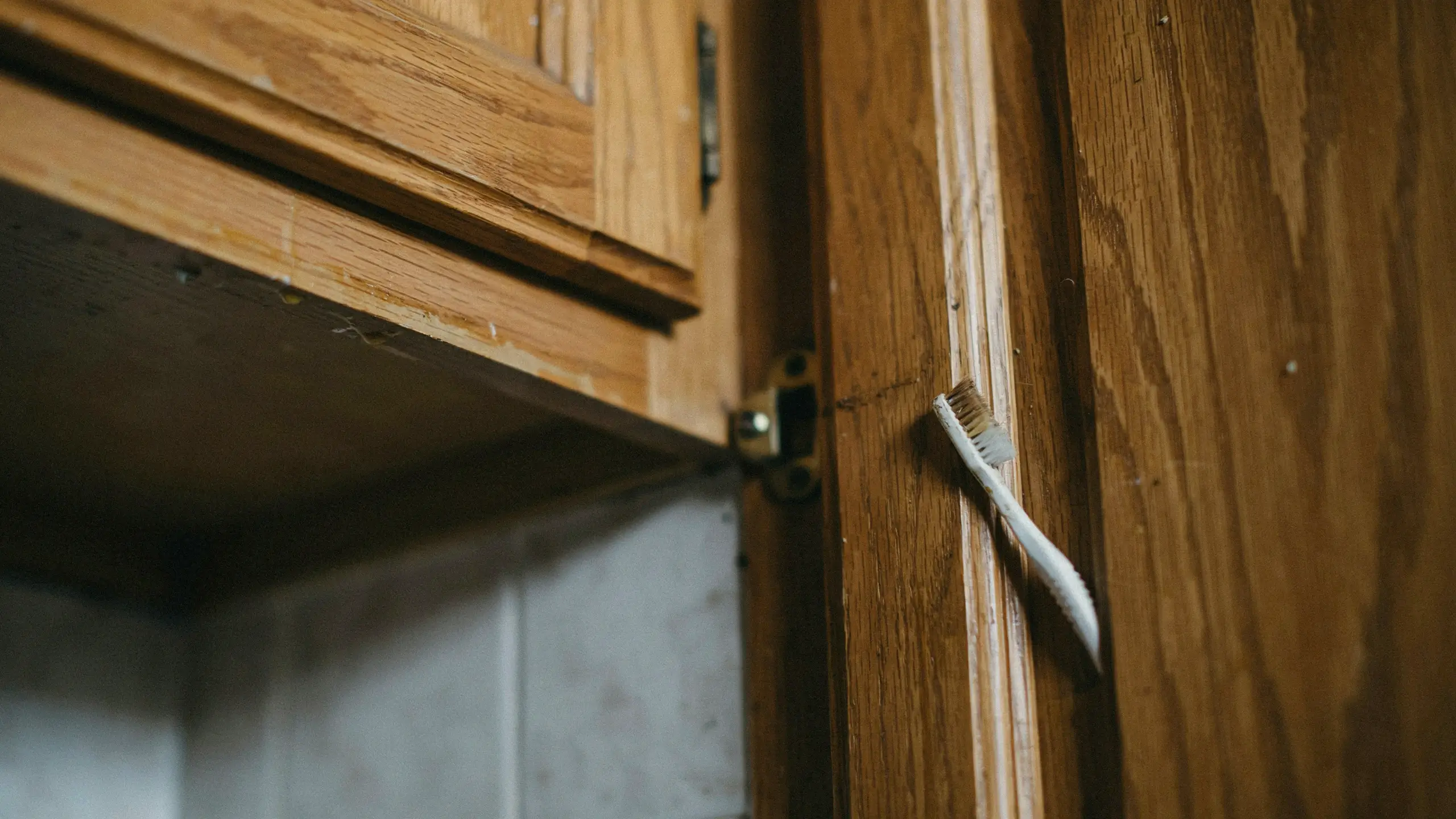 White toothbrush wedged in the narrow crevice of a wooden bathroom cabinet door, illustrating a practical cleaning hack for tackling overlooked surfaces, hard-to-reach gaps, and forgotten home maintenance areas.