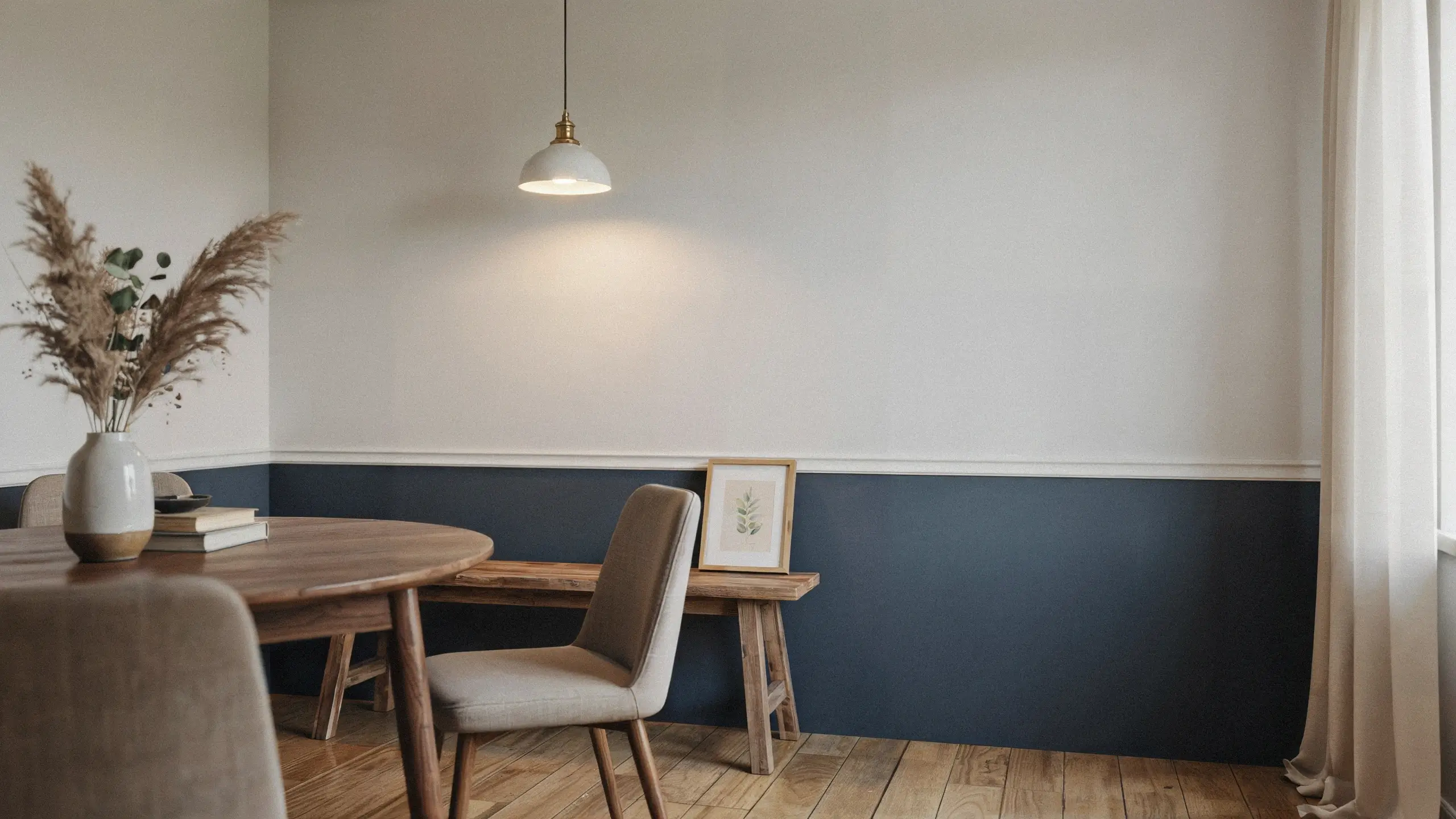 Minimalist dining room with a two-tone wall and painted wainscoting line, featuring a wooden table, pendant light, and pampas grass arrangement. 