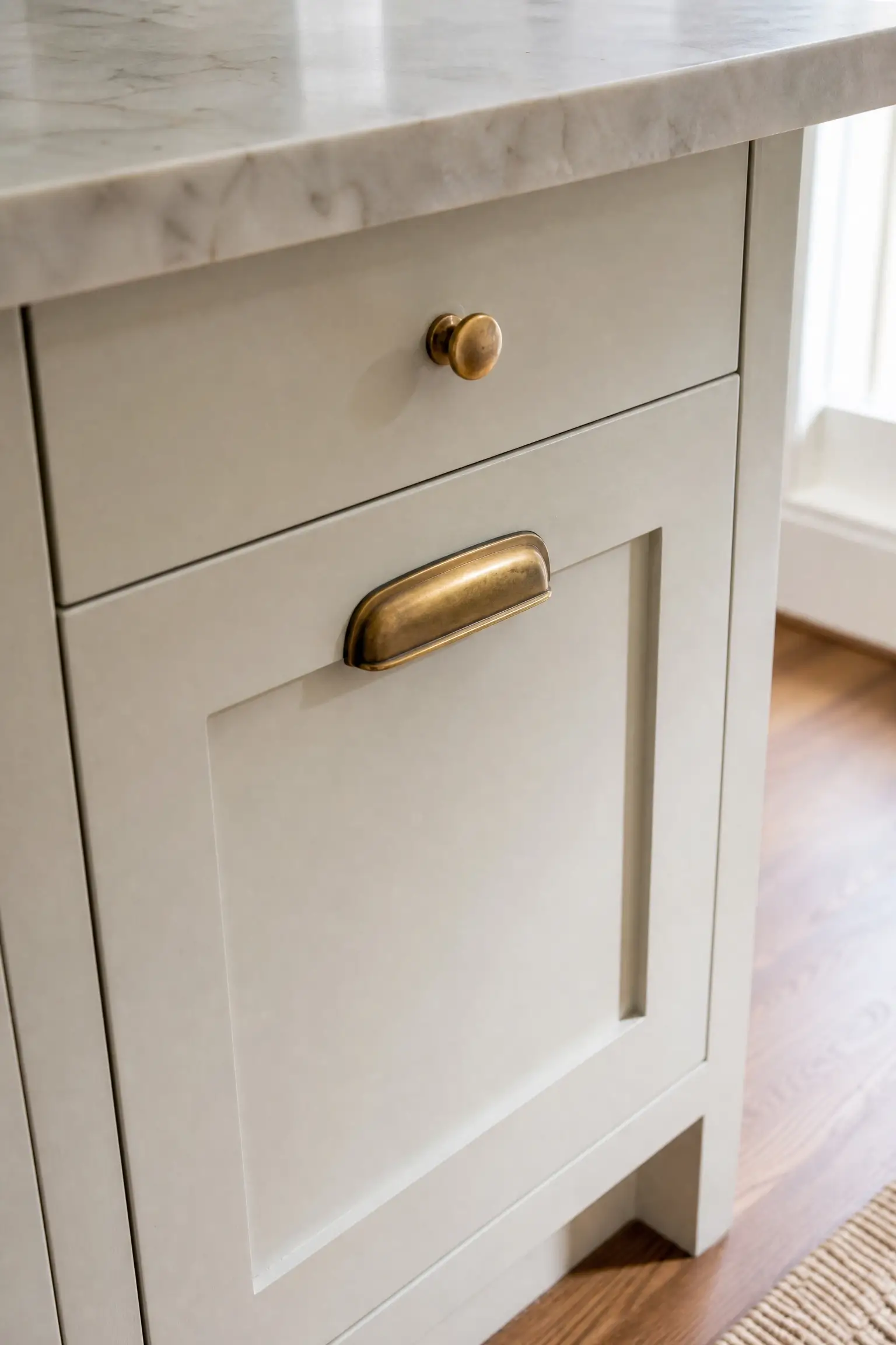 Unlacquered brass knob and bin pull on a cream shaker kitchen cabinet with marble countertop.